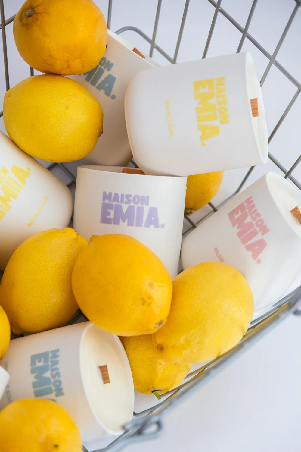 Citrons jaunes et mugs blancs avec le texte 'Maison Emia' dans différentes couleurs, dans un panier en métal.