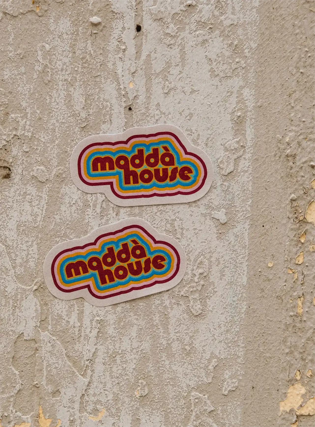 Deux autocollants colorés avec l'inscription "modda house" collés sur un mur en béton blanc.