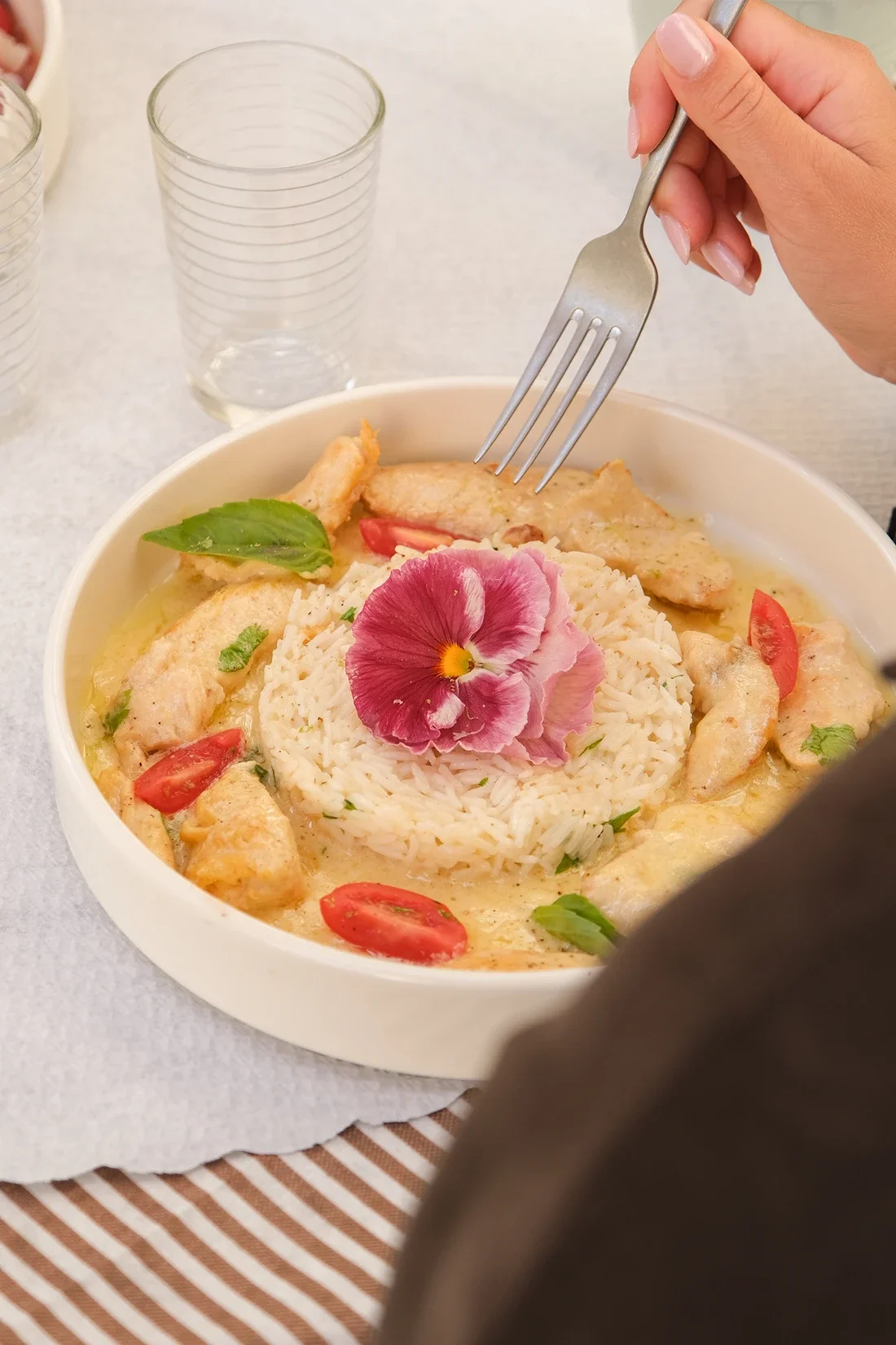Assiette de poulet en sauce avec du riz, garnie de fleurs comestibles et de tomates cerises, accompagnée d'un verre d'eau.