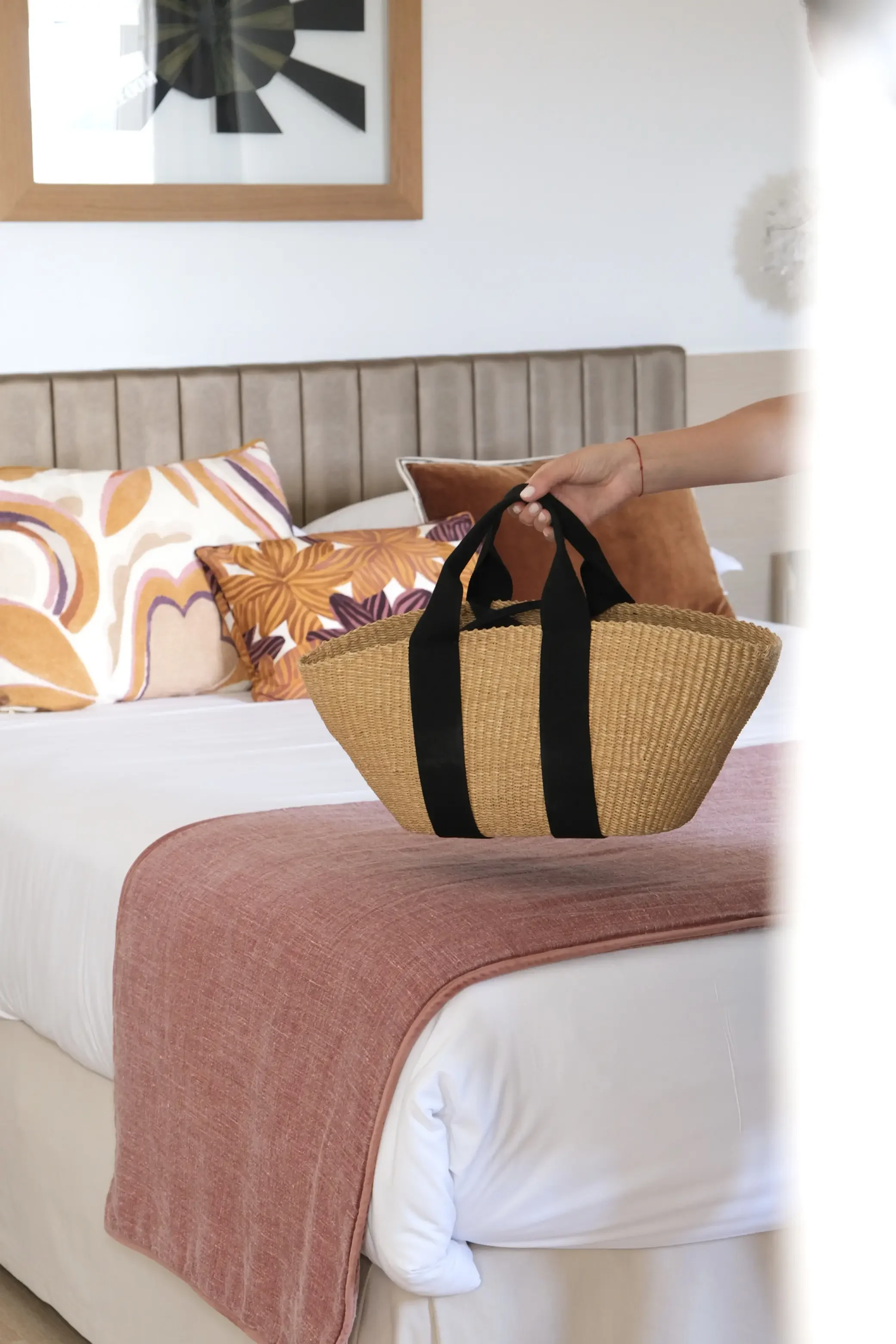 Une main tenant un sac en osier avec des poignées en tissus noirs, posé sur un lit avec des draps blancs et une couverture rose. Sur le lit, il y a deux coussins, un orange et un à motif floral.