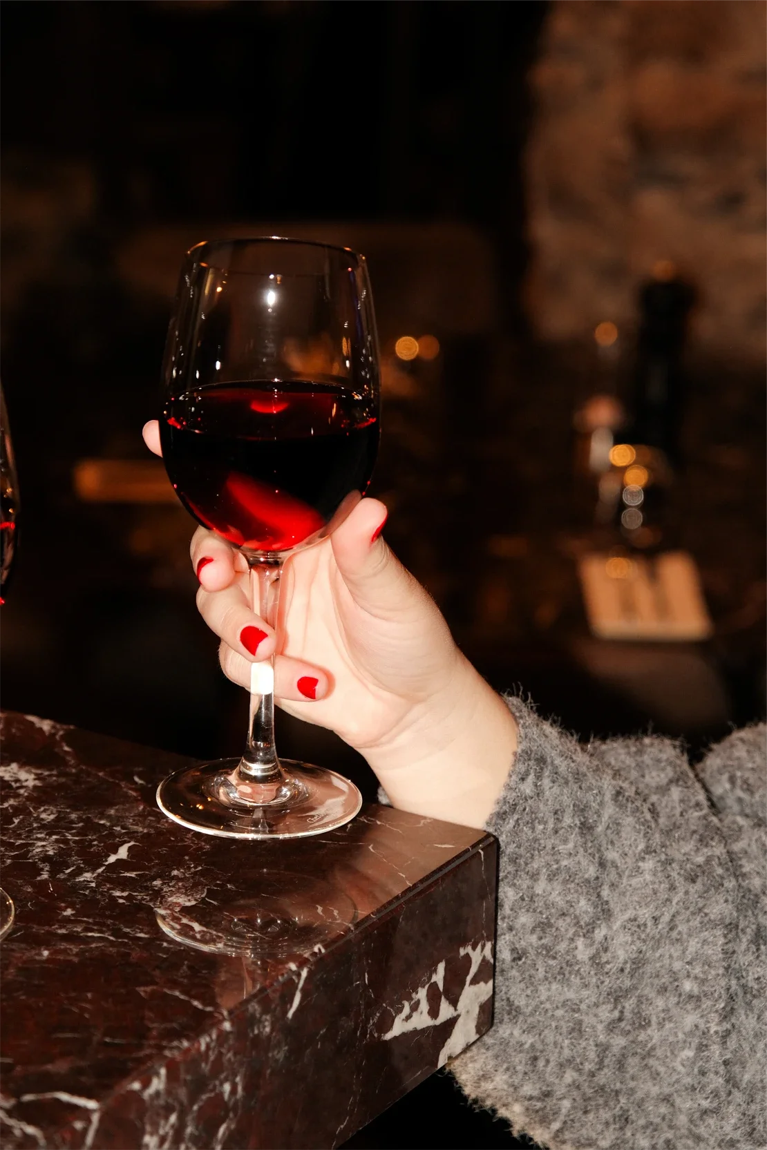 Main tenant un verre de vin rouge tenu par une personne avec des ongles peints en rouge, dans un environnement sombre avec un arrière-plan flou.