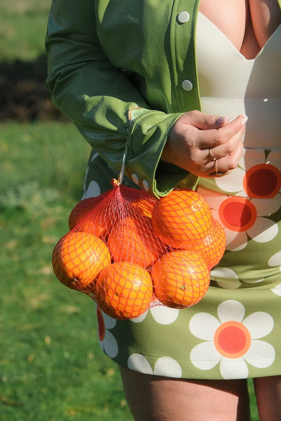 Une personne portant une robe à motifs de fleurs blanches et oranges, tenant un sac en filet rouge rempli d'oranges, avec une veste verte.