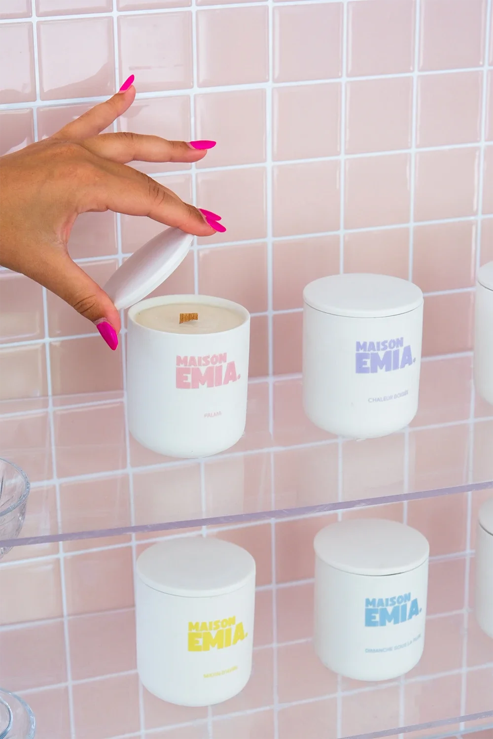 Une main avec des ongles rose vif ouvre un candle dans une salle de bain avec un mur en carreaux roses. Sur la paroi, quatre bougies de différentes couleurs et noms sont visibles.