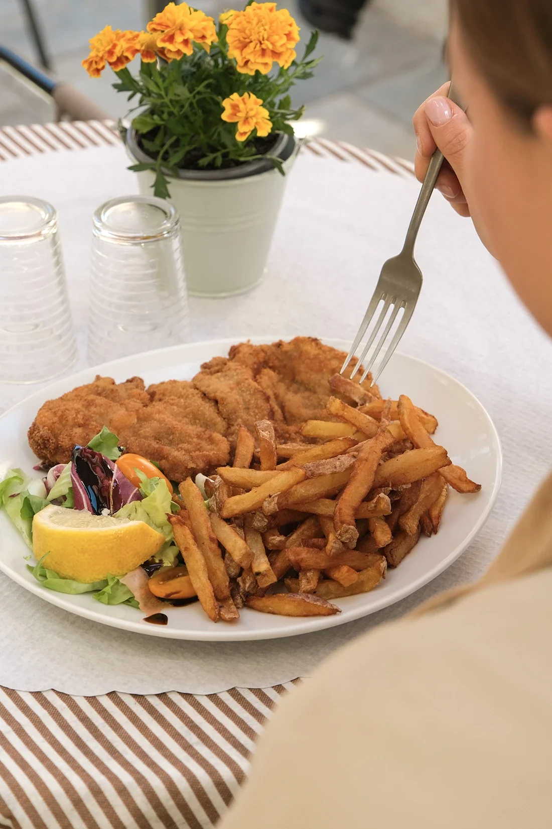 Assiette de nourriture comprenant du poulet frit, des frites, une salade avec un quartier de citron, et des tomates cerises. En arrière-plan, une plante en pot avec des fleurs jaunes ou orange, et trois verres en plastique empilés.