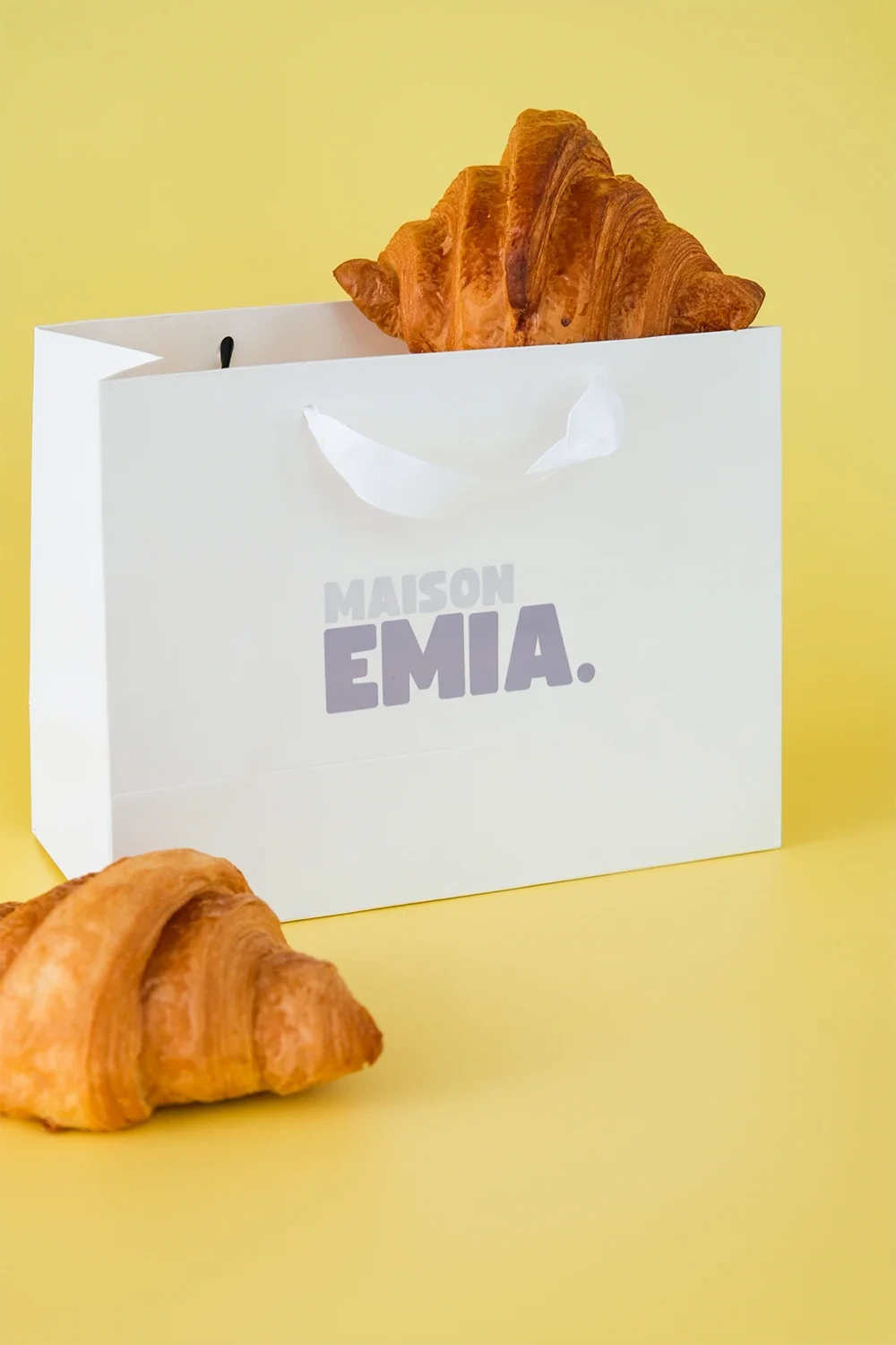 Deux croissants dans et à côté d'une boîte blanche avec l'inscription "MAISON EMIA" sur fond jaune.