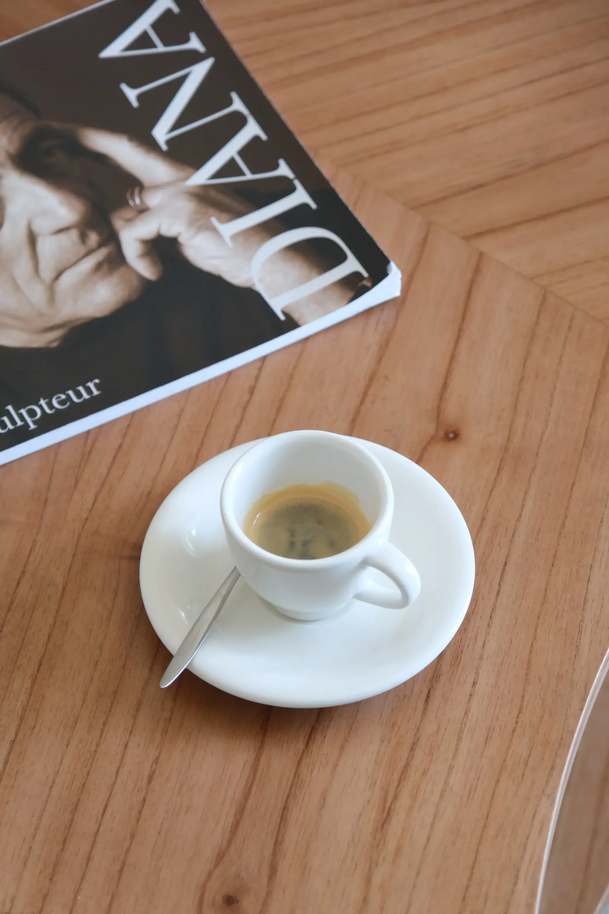 Une tasse de café servie dans une petite tasse blanche sur une assiette blanche, avec une cuillère en métal. La tasse est posée sur une table en bois.
