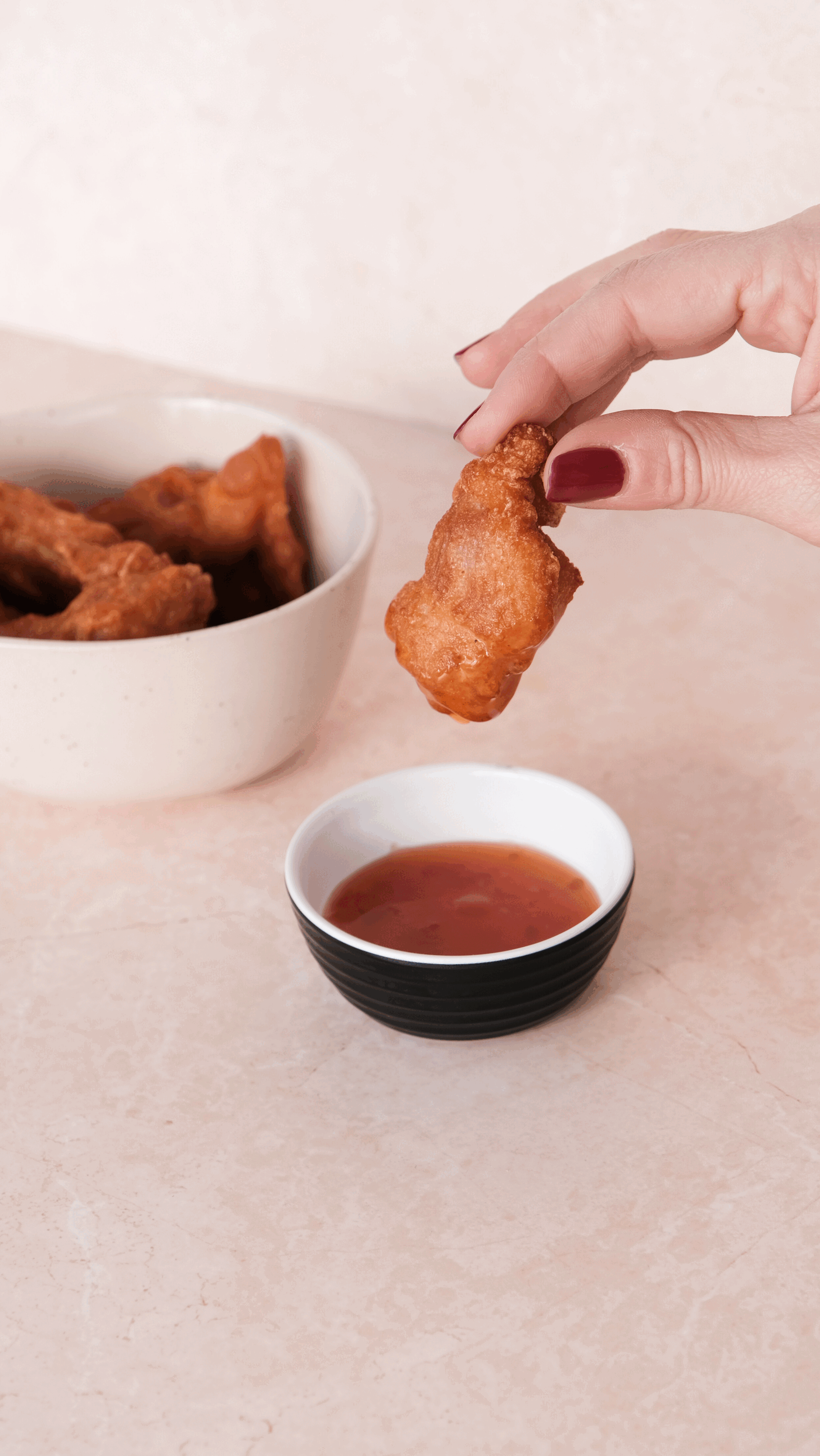 Une main tenant un morceau de poulet frit au-dessus d'une sauce rouge dans un bol noir, avec un bol blanc de sauce à coté et un bol blanc de morceaux de poulet frits en arrière-plan.
