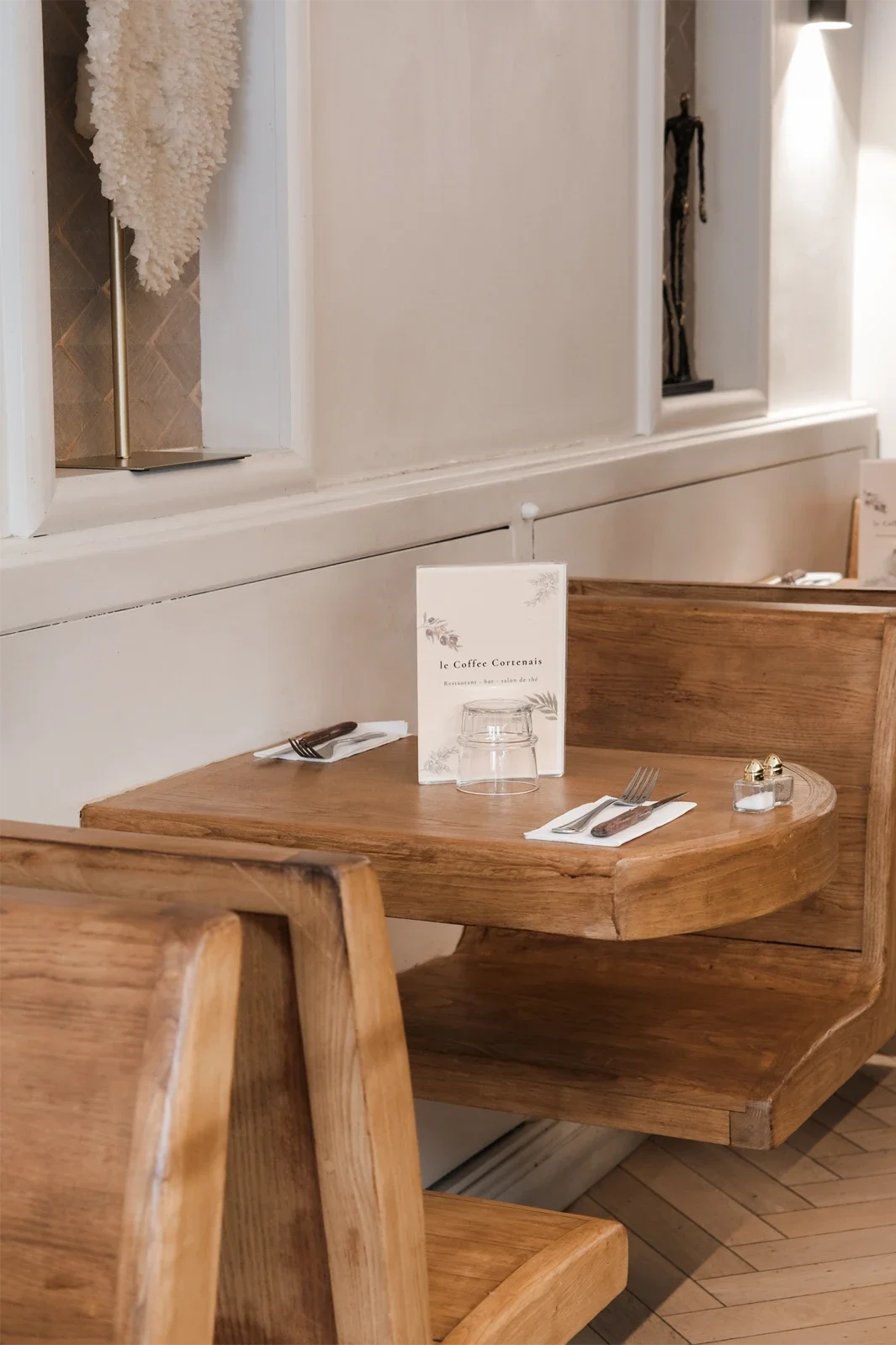 Une table en bois dans un restaurant, avec un menu, une verre à boire, des couverts et des assaisonnements.