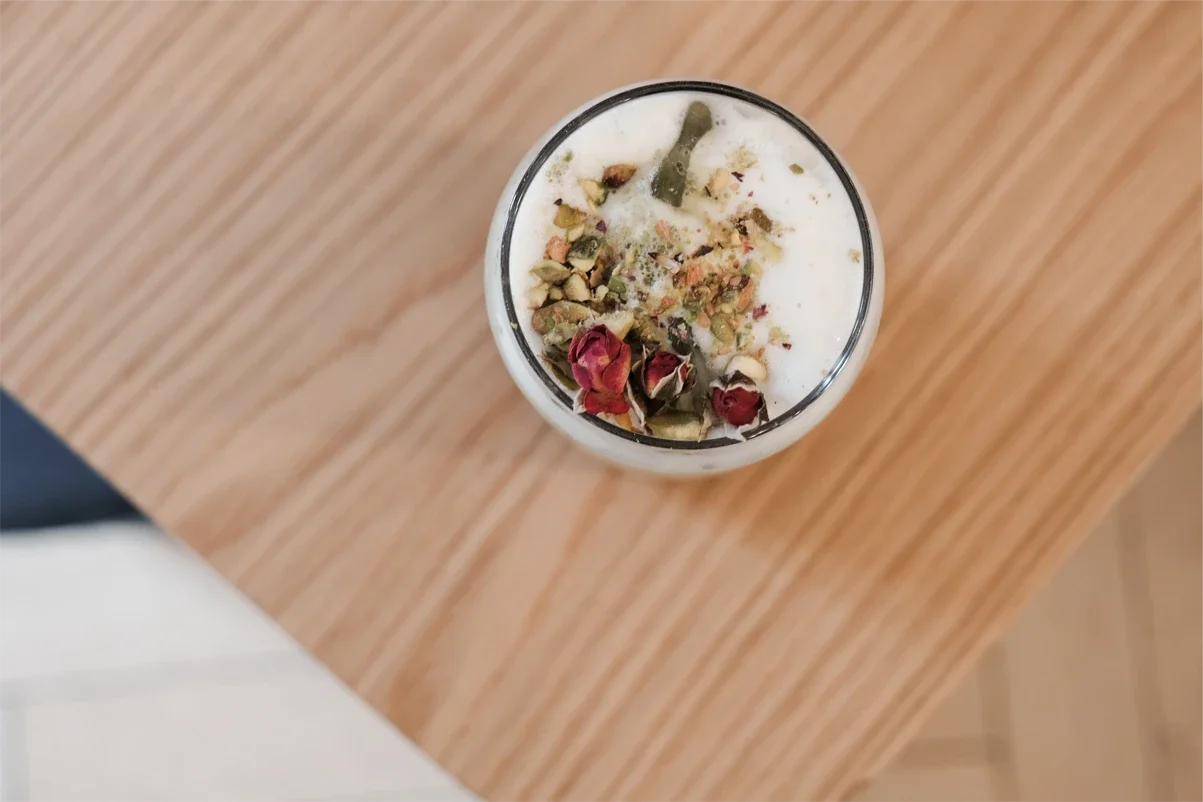 Verre de lait chaud garni de roses séchées et de pistaches concassées, posé sur une table en bois.