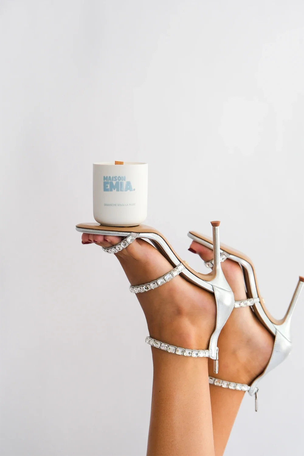 Une main portant une paire de talons hauts en blanc avec des détails en strass, tenant un porte-bougie blanc orné de texte bleu et gris.