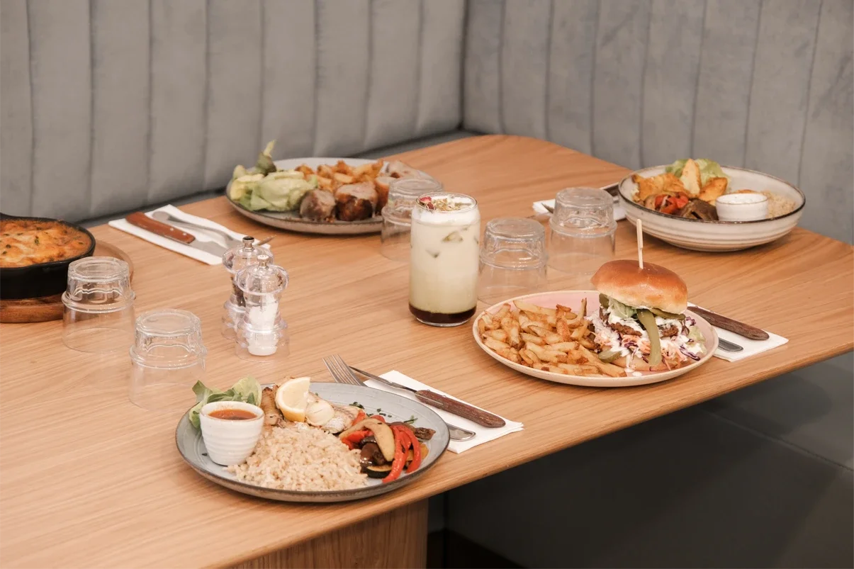 Repas varié sur une table en bois avec des assiettes de nourriture, des verres, une boisson au lait, un burger avec des frites, un plat de riz avec légumes et de la sauce. Arrière-plan en mur en bois clair.