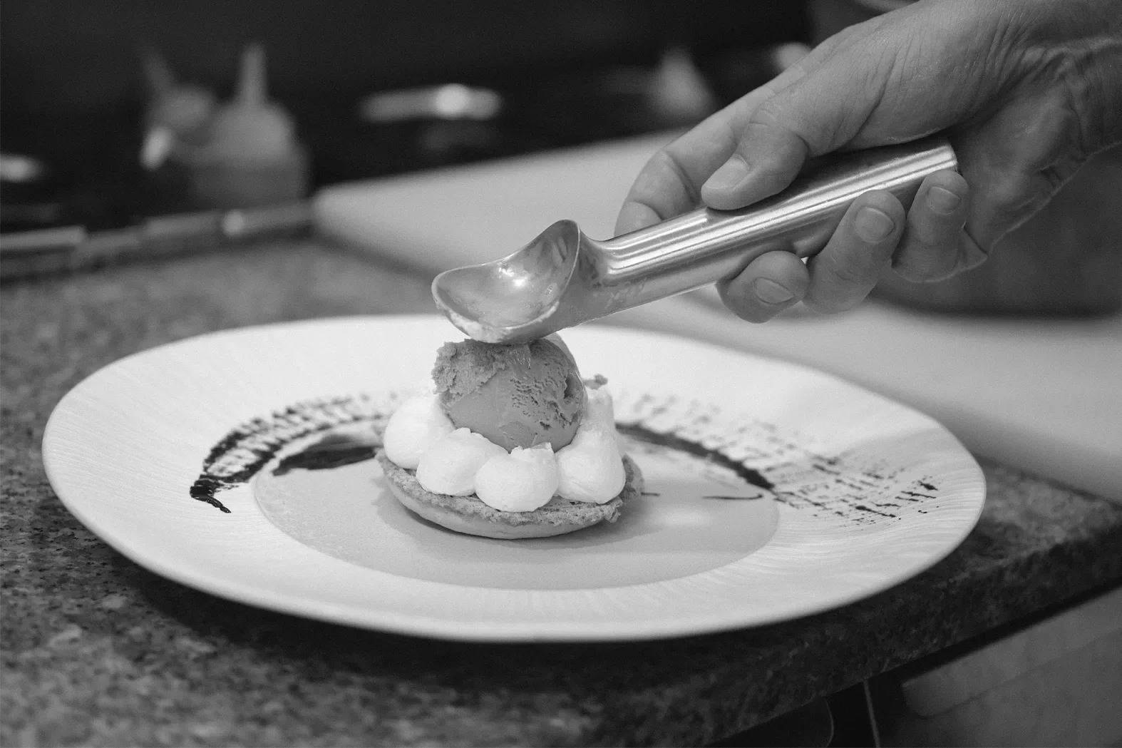 Une main utilise une pince pour déposer une boule de glace sur une pâtisserie décorée de chantilly et de sauce chocolat sur une assiette. Arrière-plan flou, cuisine visible.