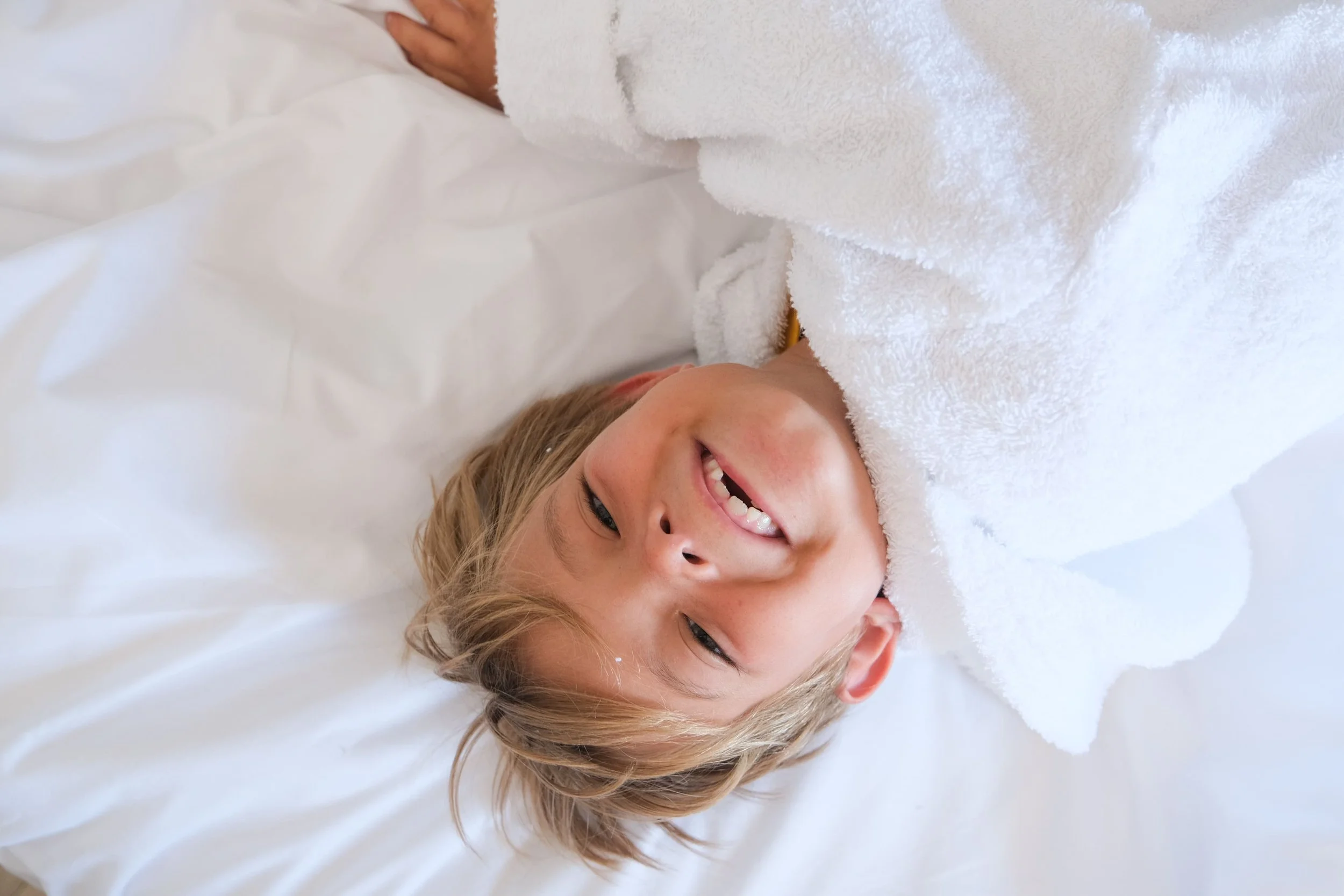 Un enfant qui sourit, allongé sur un lit, avec un peignoir blanc. 