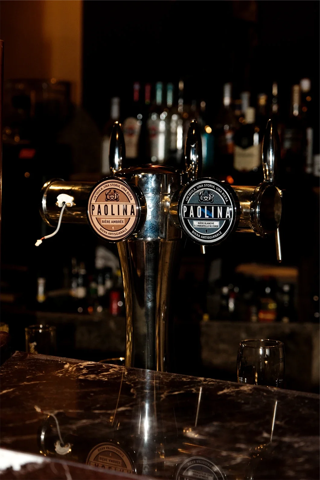 Une pompe à bière en métal avec deux robinets, l'un pour une bière ambrée et l'autre pour une bière blanche, dans un bar. En arrière-plan, des bouteilles d'alcool sur une étagère.