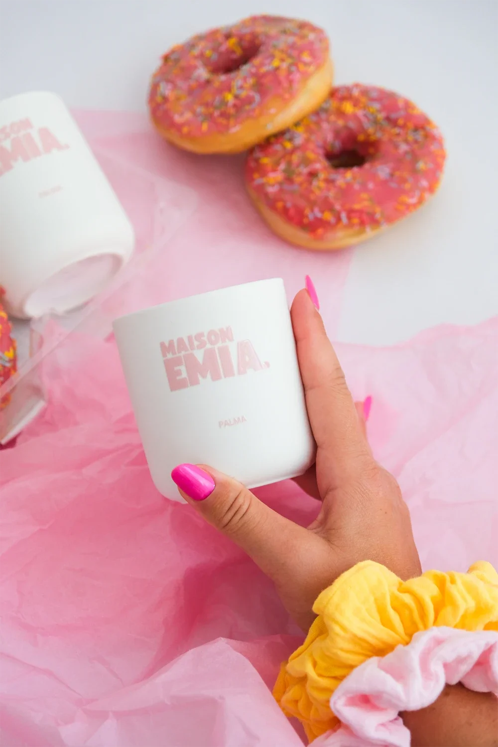 Une main tenant une tasse blanche avec le texte « MAISON EMIA » écrit en rose pâle, entourée de donuts roses avec des sprinkles, sur un fond rose, et une autre tasse blanche similaire à proximité.