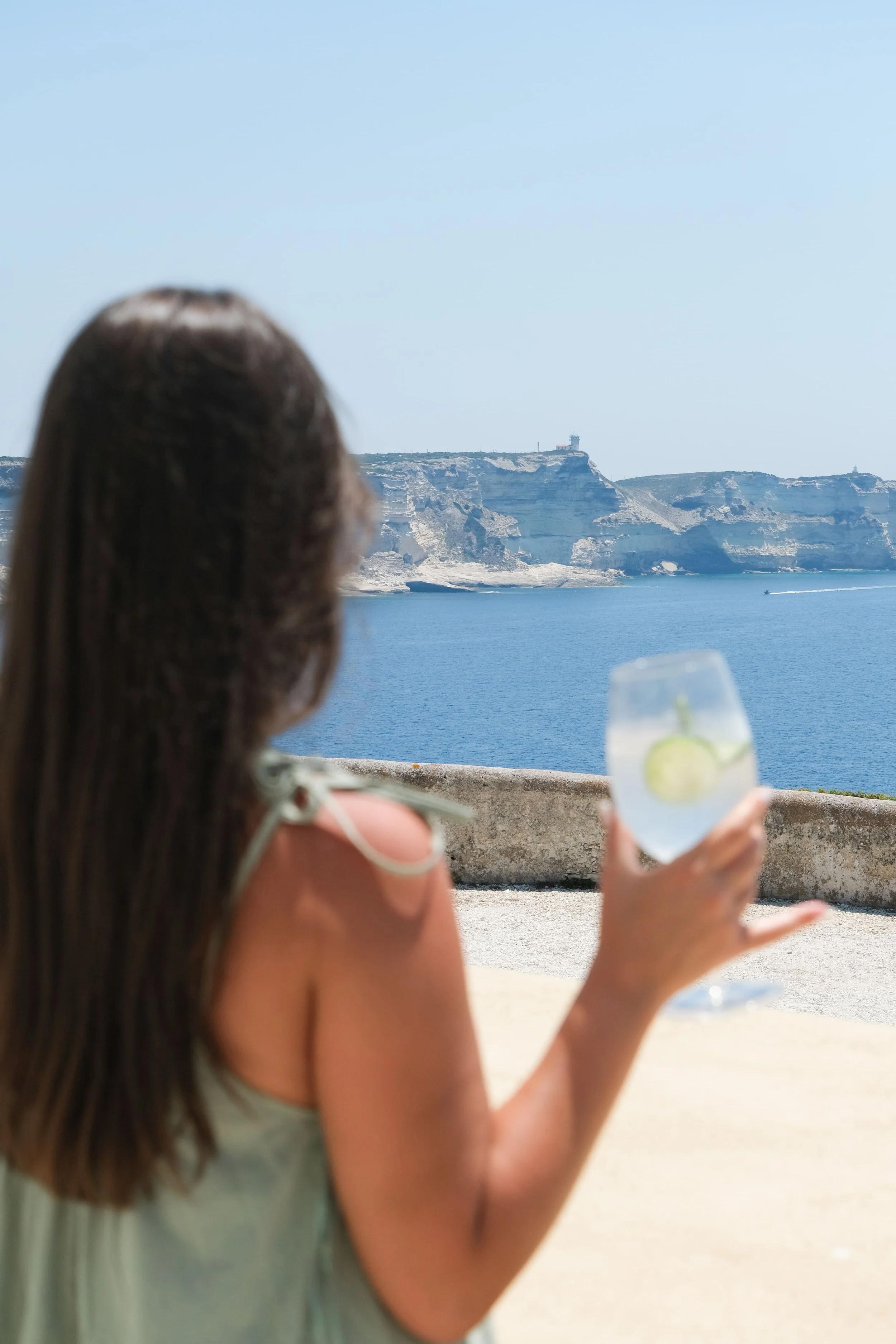 Une personne de dos qui tient une boisson dans la main et qui regarde la vue sur la mer, et les falaises.