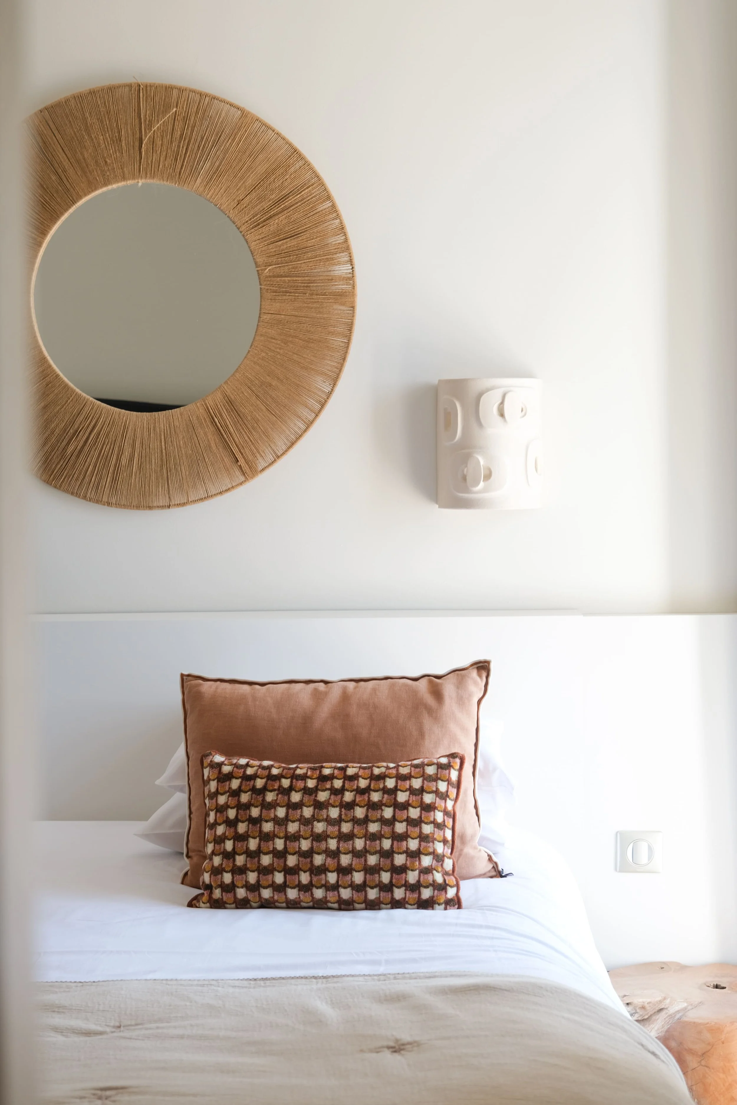 Une chambre d'hôtel avec un lit, une table de nuit en bois à droite, une applique et un miroir au dessus du lit. Sur le lit, un dessus de lit beige et trois coussins.