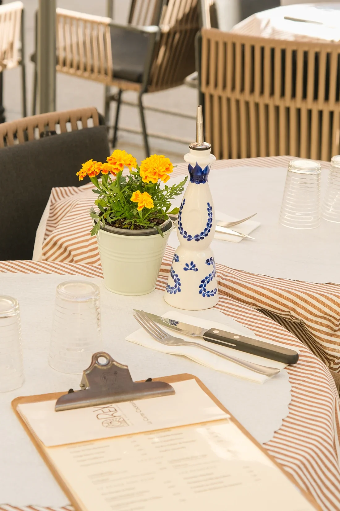 Une table de restaurant avec un pot de fleurs contenant des fleurs jaunes, un objet à vaporiser décoratif blanc et bleu, des verres en plastique inversés, des couverts placés sur une serviette blanche, un menu, une planche en bois et un carnet avec u