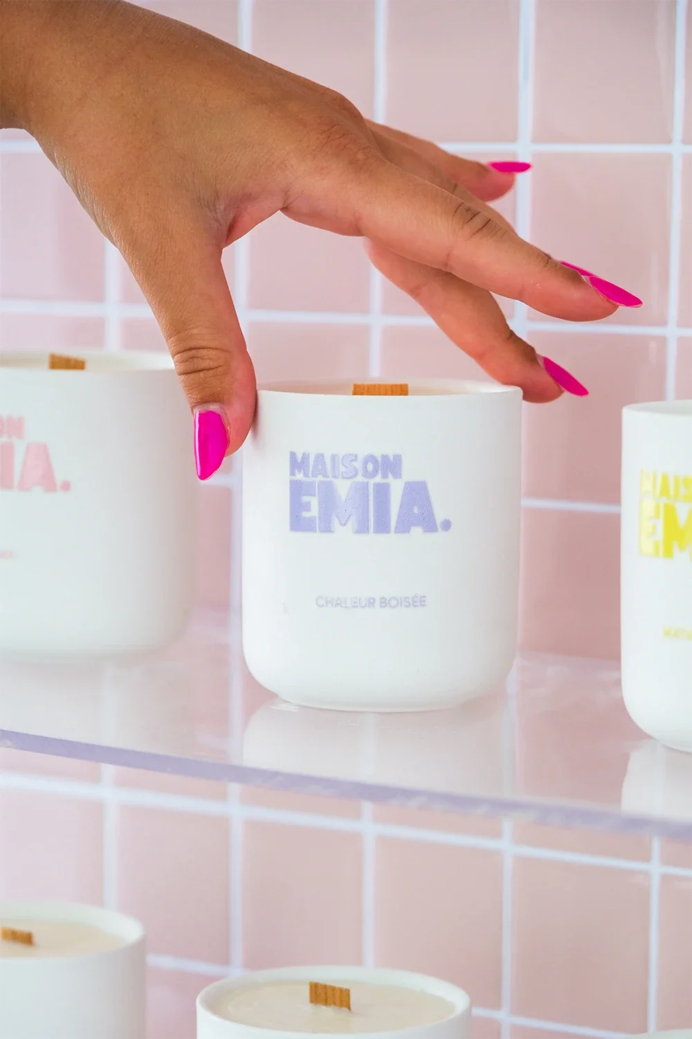 Bougie parfumée blanche avec l'inscription "MAISON EMIA" en violet, sur une étagère avec un fond de carreaux roses.