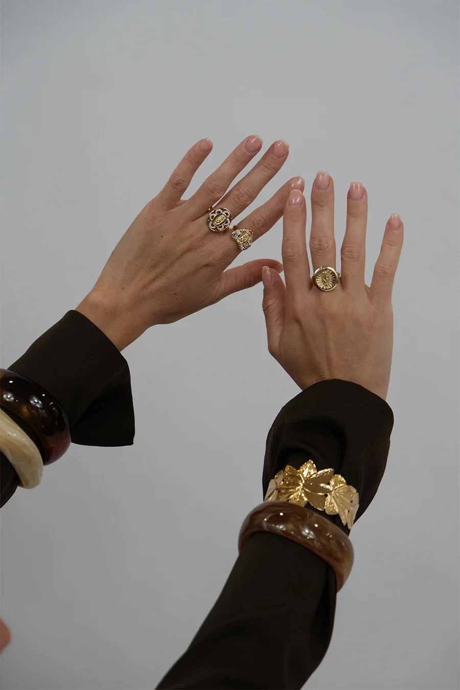 Deux mains avec plusieurs bagues en or et bijoux. Mains portant aussi des bracelets en résine ou en pierre. Fond gris.