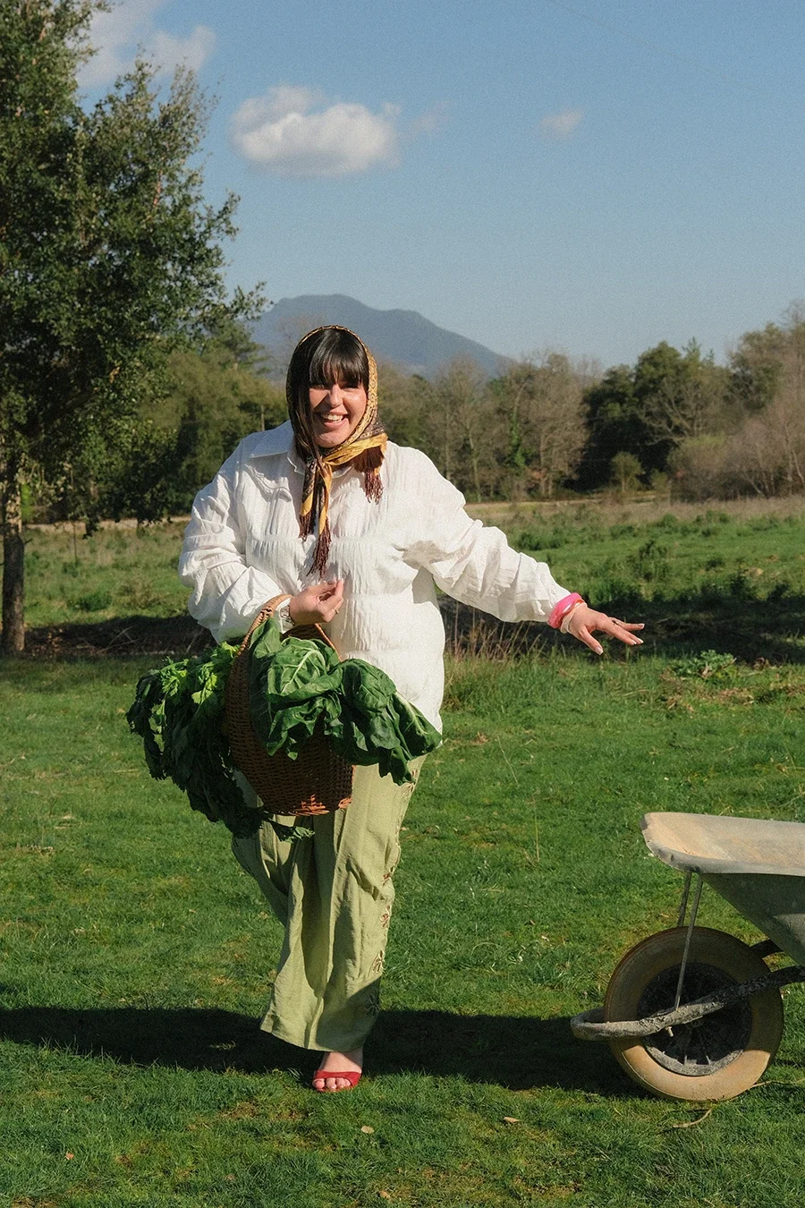 Femme souriante portant un panier de légumes verts, debout dans un champ ensoleillé avec des arbres et des montagnes en arrière-plan.