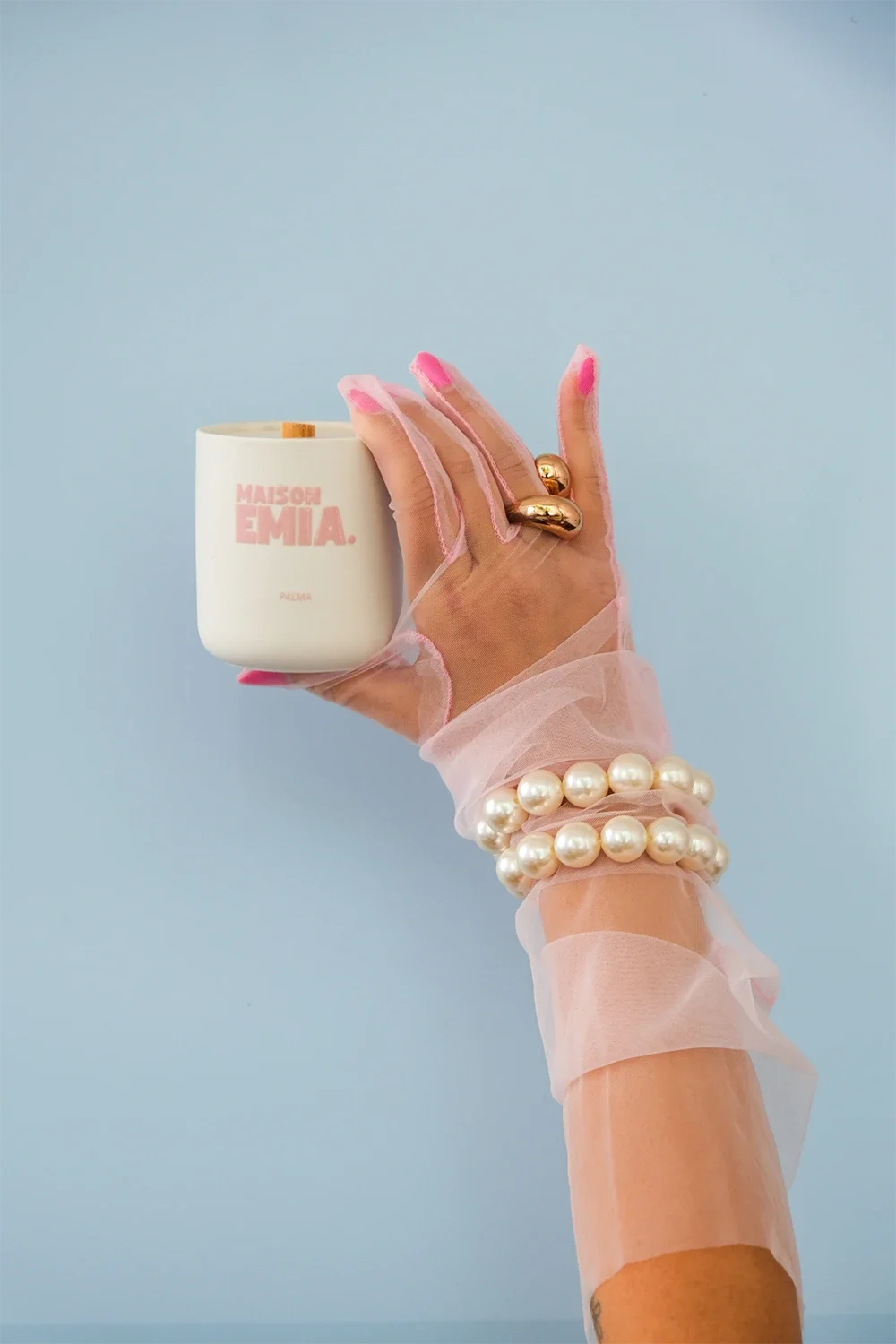 Une main féminine portant des bijoux perles et un gant en tulle rose, tenant une bougie blanche avec l inscription "MAISON EMIA" sur un fond bleu clair.