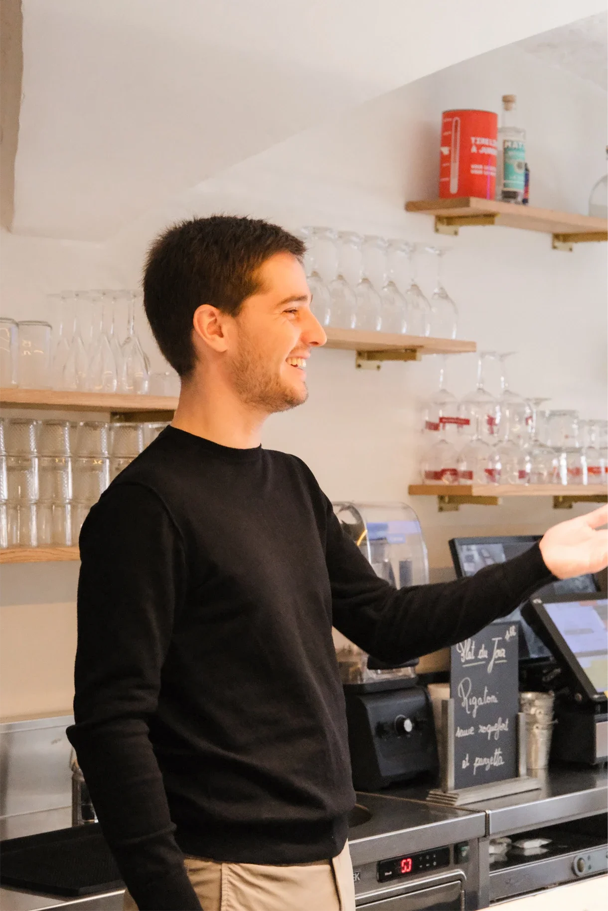 Homme souriant portant un pull noir dans un café ou restaurant, avec des étagères contenant des verres et des bouteilles en arrière-plan, et un écran de caisse.