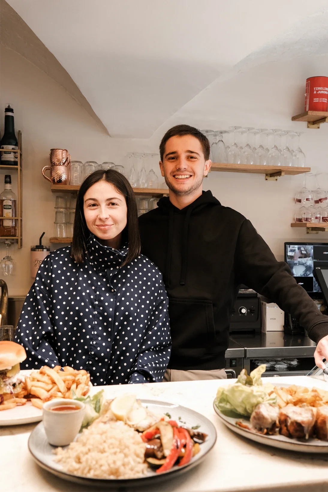 Deux personnes, un homme et une femme, souriant derrière un comptoir de restaurant avec des plats de nourriture en premier plan.