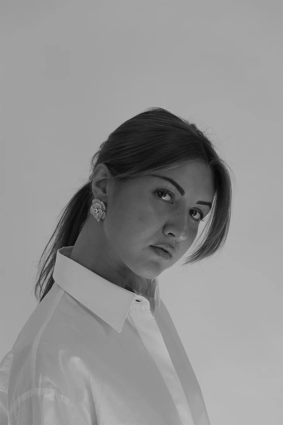 Portrait en noir et blanc d'une femme avec des cheveux bruns attachés, portant des boucles d'oreilles et une chemise blanche, regardant vers la caméra