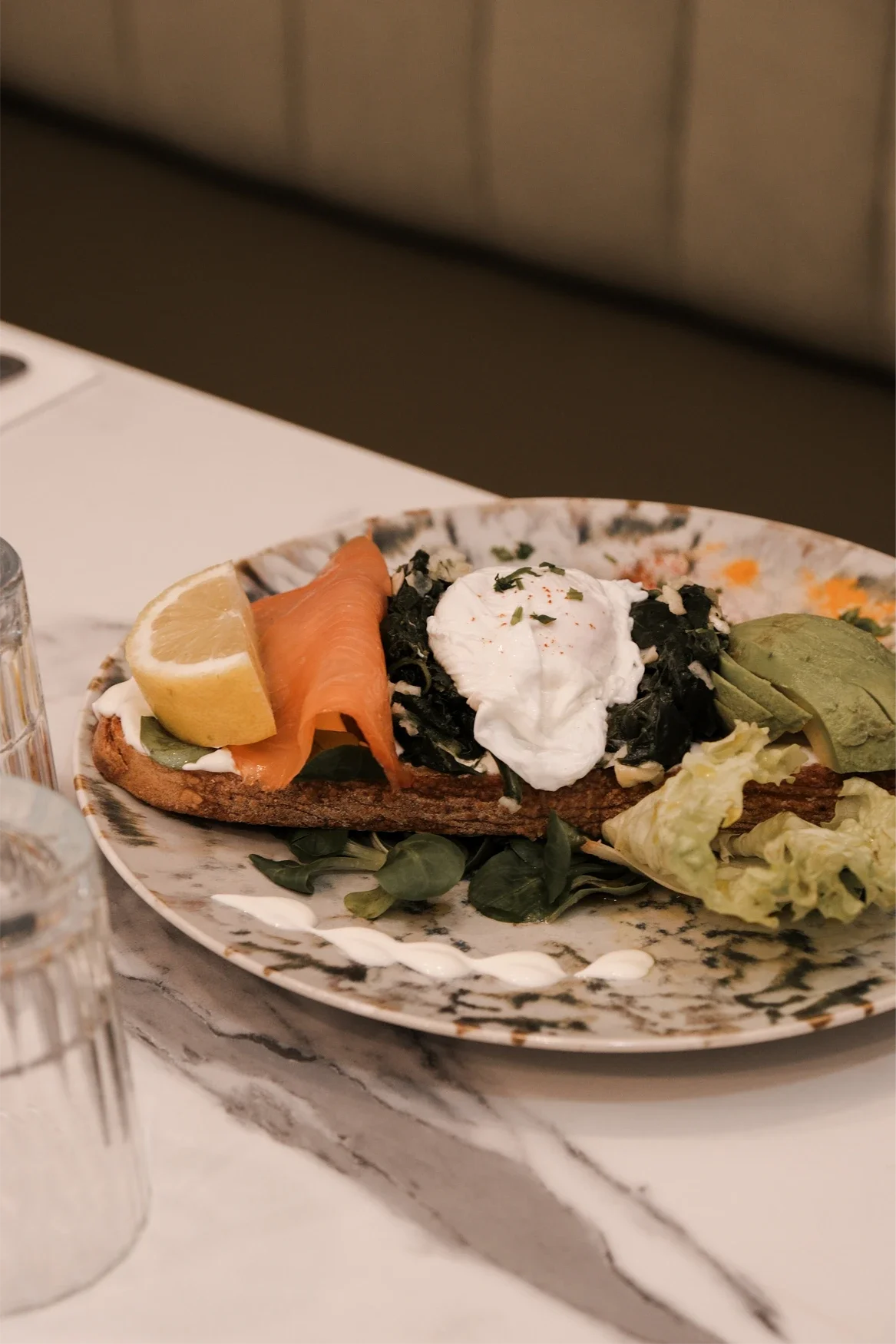 Une assiette avec du pain, du saumon fumé, un demi-citron, du fromage avocat, une salade, du fromage à la crème, des épinards en sauce et un œuf poché.