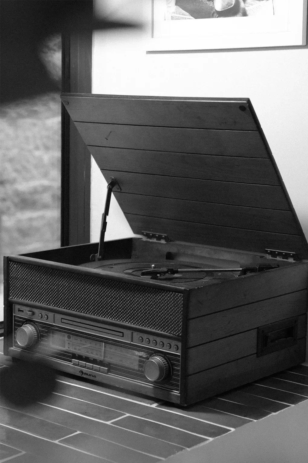 Un tourne-disque vintage en bois avec une face avant en métal et des boutons de contrôle, posé sur une table en bois, avec une photo en noir et blanc en arrière-plan.
