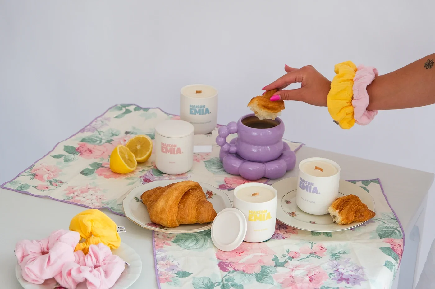 Table dressée avec des croissants, une tasse de café, des bougies, un citron coupé en deux, et des pinces à cheveux de différentes couleurs, avec une main tenant un biscuit au-dessus de la tasse de café.