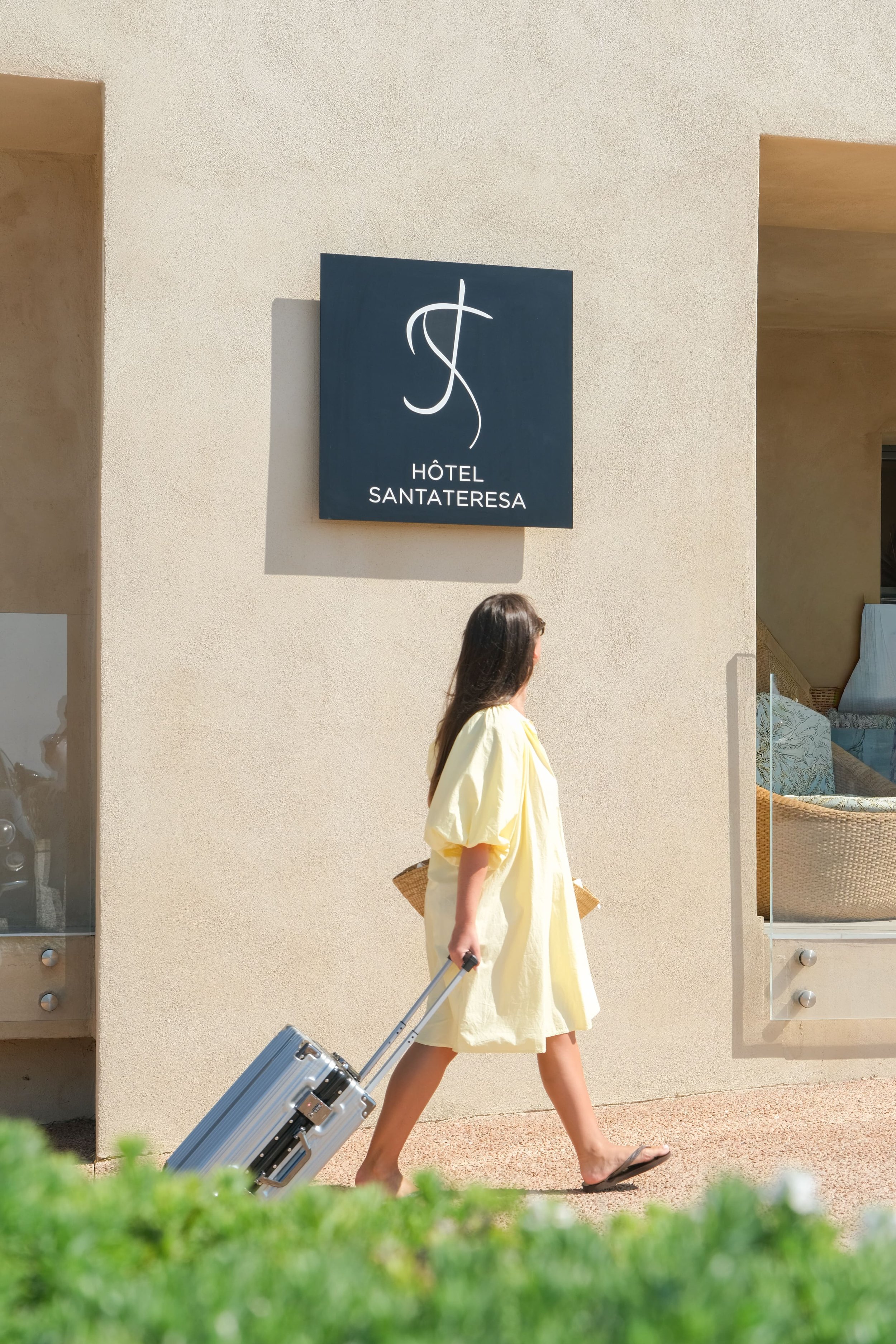 Une personne qui marche devant un hôtel, avec une robe jaune, des tongs, une valise, un panier en osier avec des poignées en tissu vert, et des lunettes.