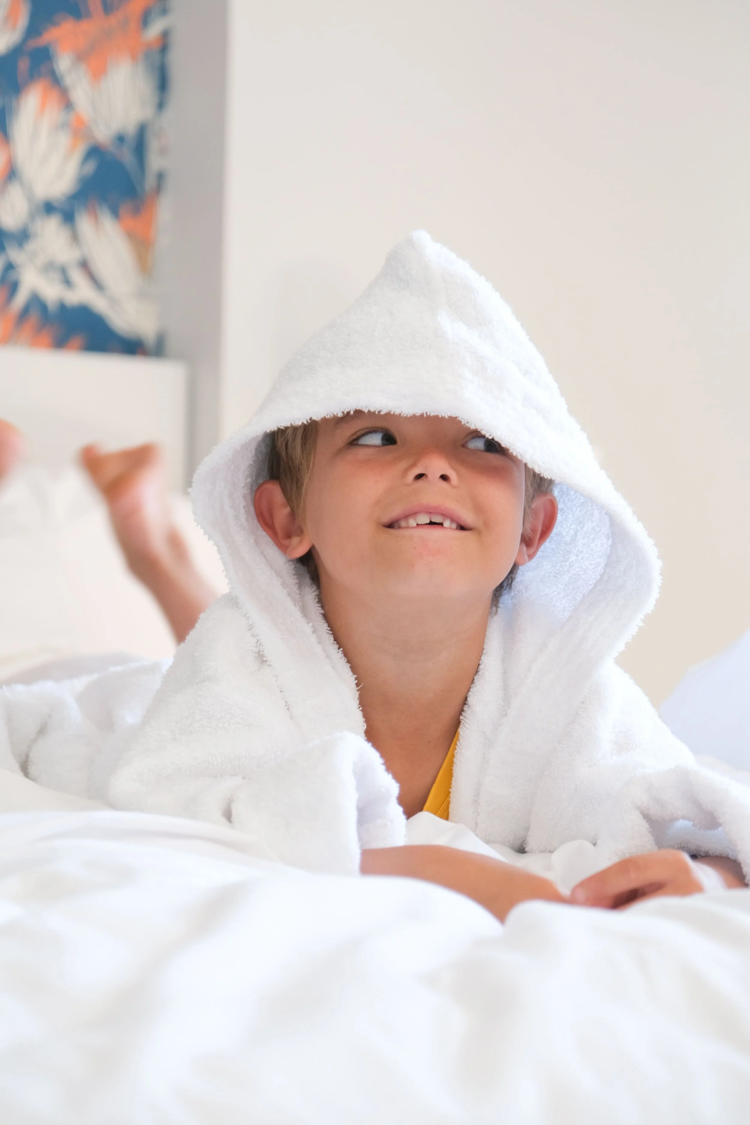 Un enfant qui regarde sur le côté, avec un peignoir blanc, sur un lit d'hôtel.