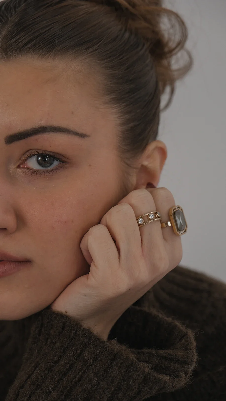 Un gros plan d'une femme avec des cheveux bruns relevés, portant plusieurs bagues, dont une avec une grande pierre grise, et une autre avec de petits diamants, posant sa main près de son visage.
