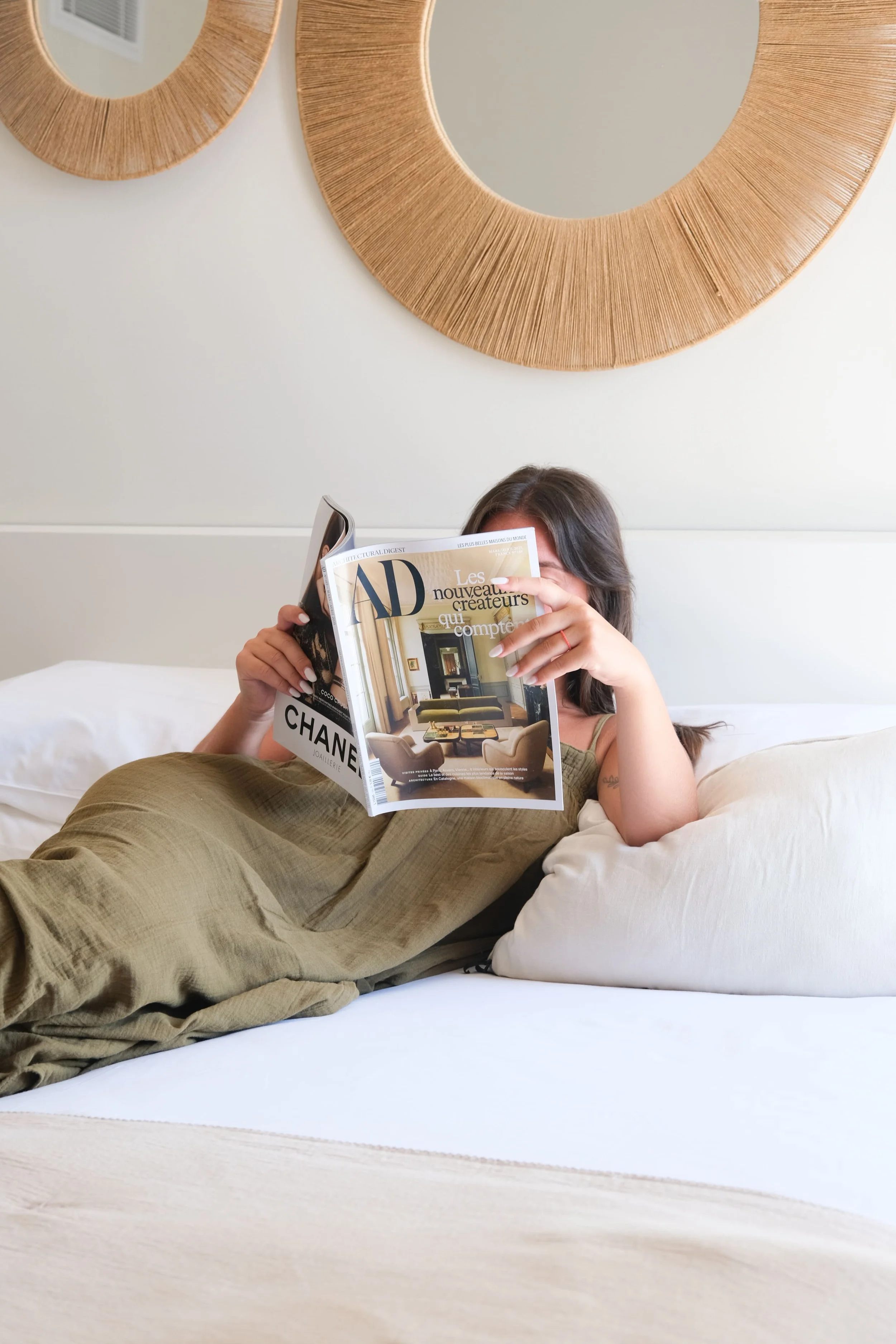 Une personne allongée sur un lit d'hôtel, la personne porte une robe kaki et lit un magazine. Sur le lit se trouve des coussins et un dessus de lit beige. Au dessus du lit deux miroirs.