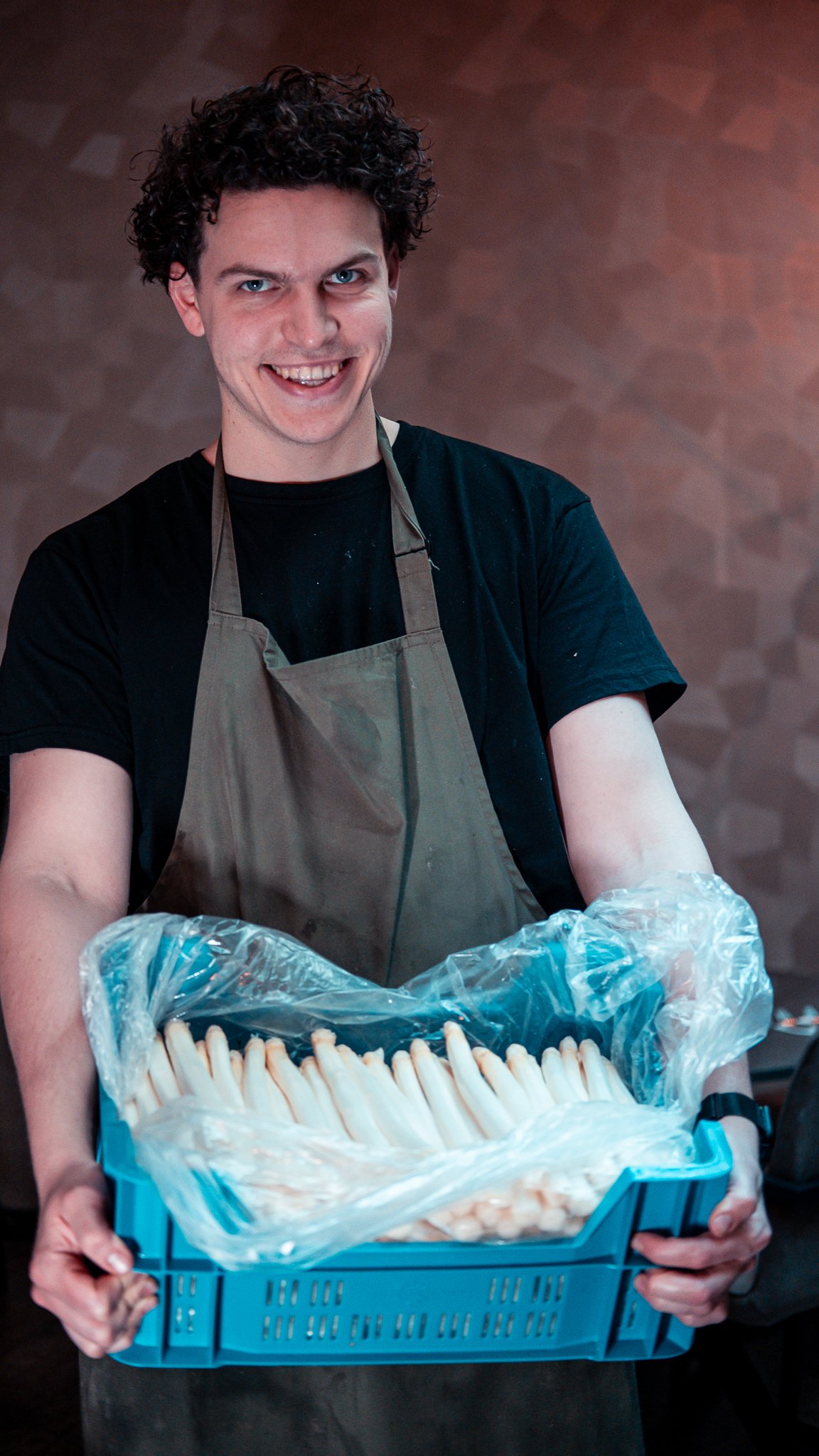 Smiling man wearing an apron and holding a blue crate filled with white asparagus.