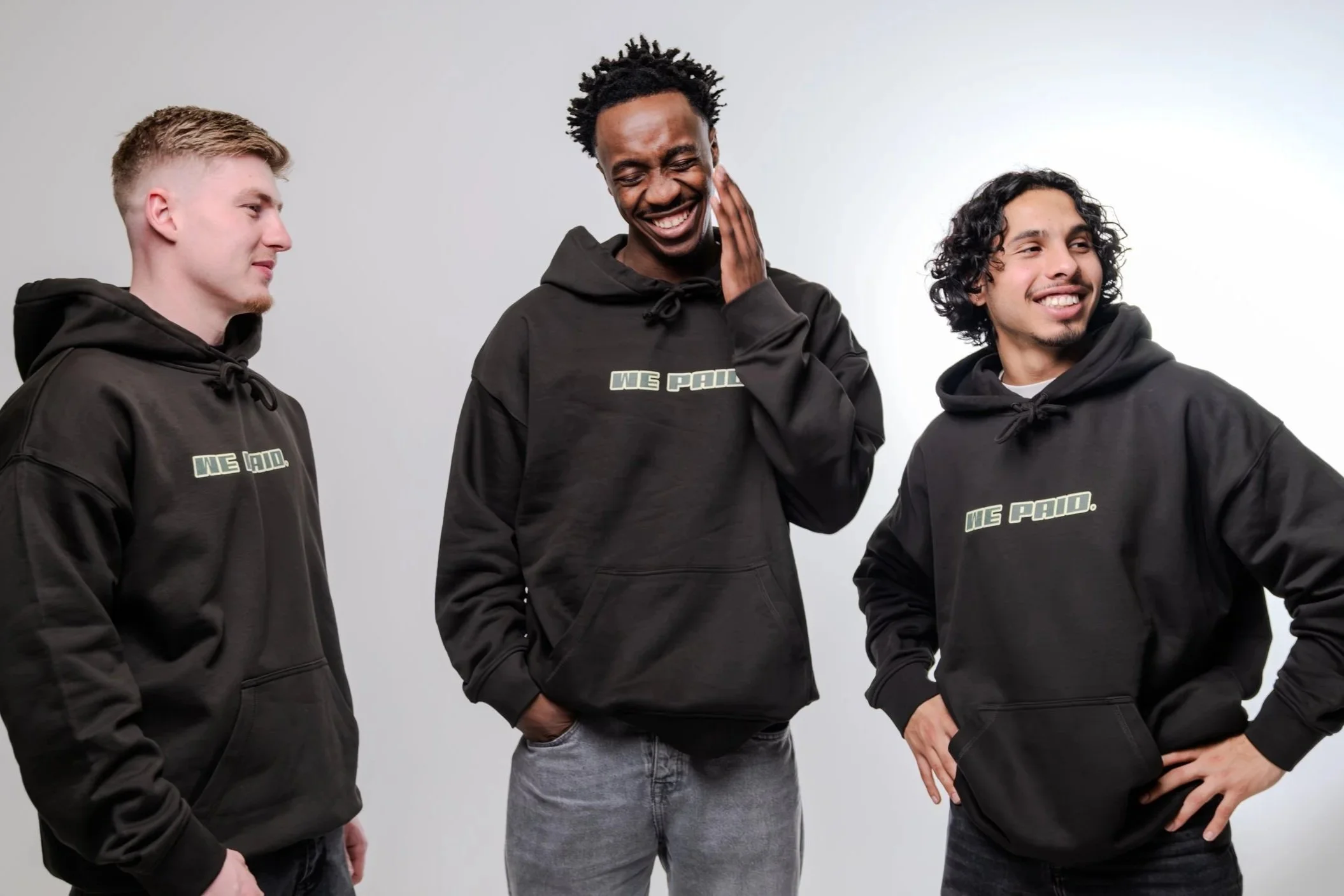 Drietal jongens in zwarte hoodies met tekst 'WE PAID' die lachen en poseren tegen een witte achtergrond.