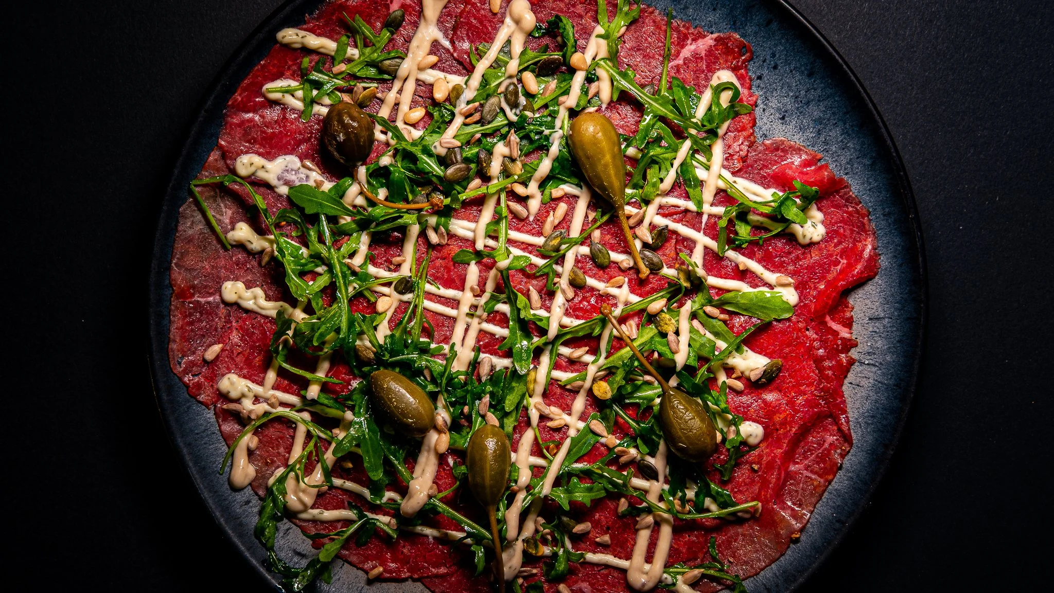 Carpaccio topped with rocket, caper berries, pine nuts and creamy dressing on a dark plate.