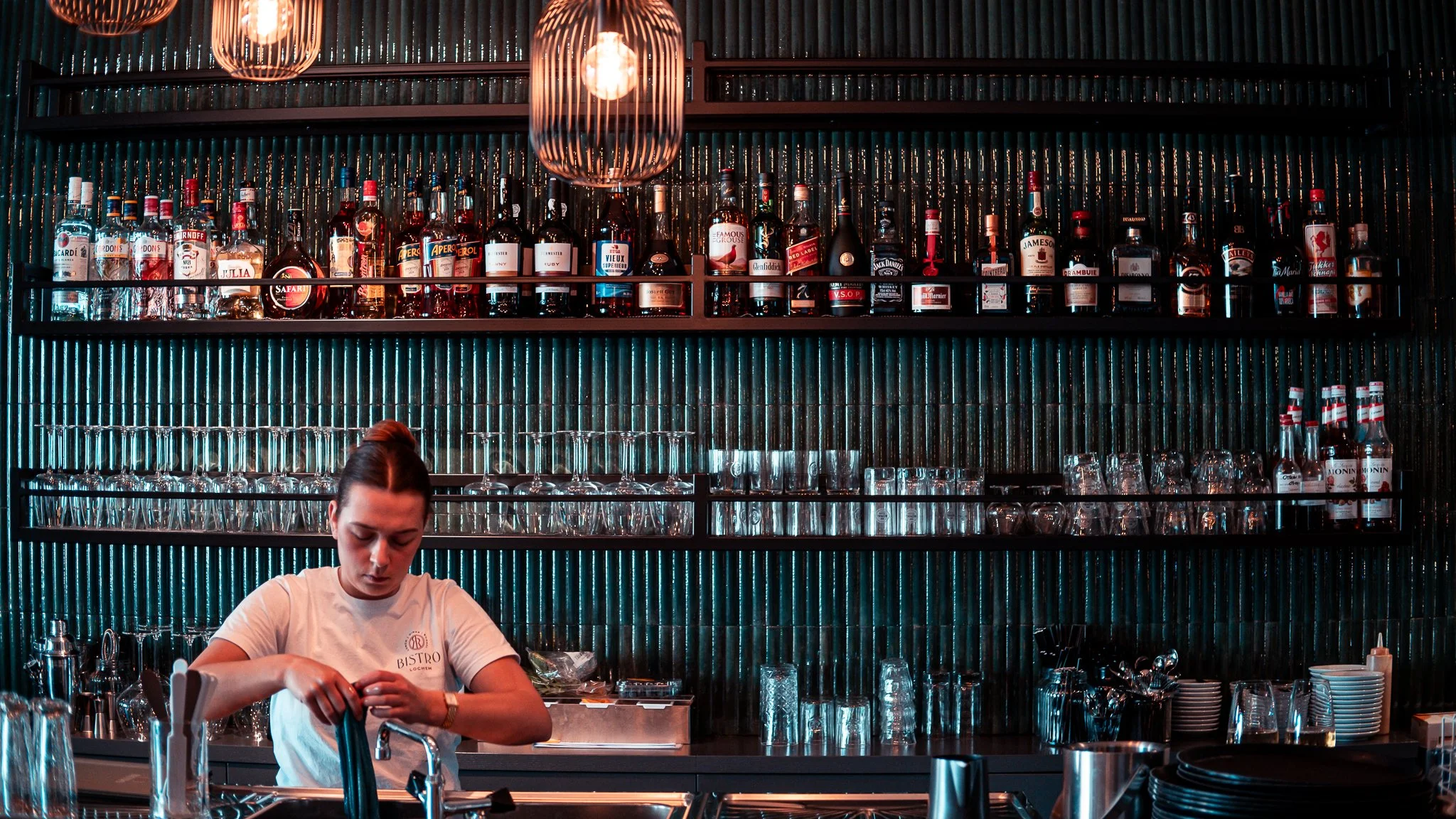 Staff member behind a restaurant bar, with shelves of bottles and glassware beneath warm pendant lights.