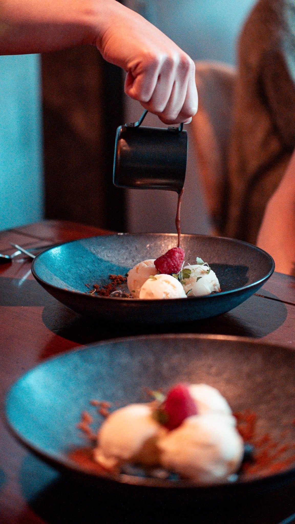 Hand pouring sauce from a black jug over a dessert topped with a raspberry in a dark bowl.