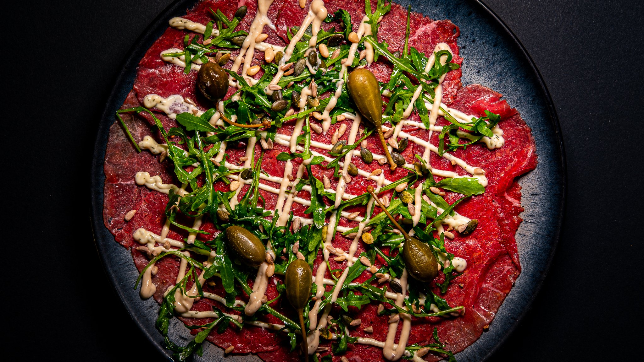 Carpaccio topped with rocket, caper berries, pine nuts and creamy dressing on a dark plate.