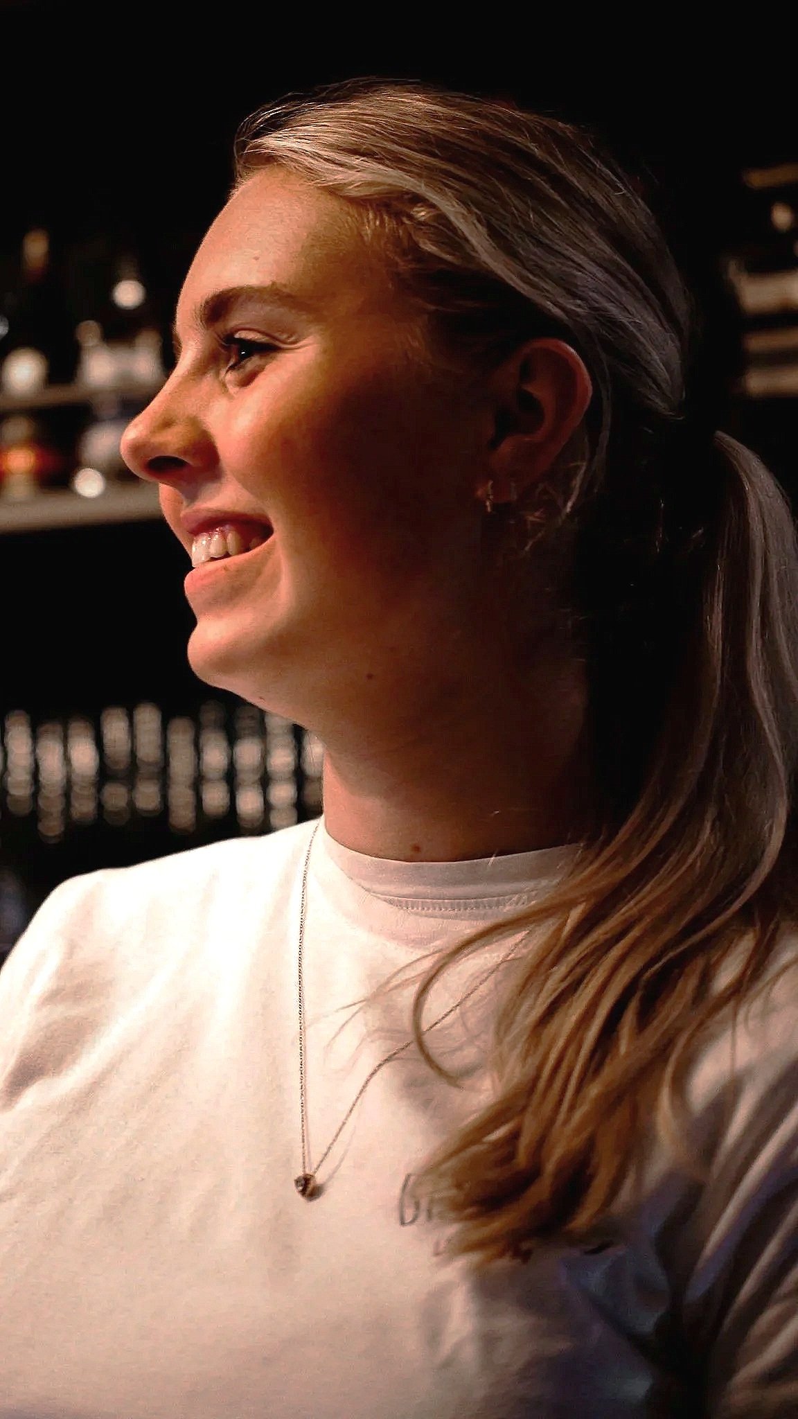 Smiling woman with a ponytail in profile, with softly blurred bar shelves in the background.