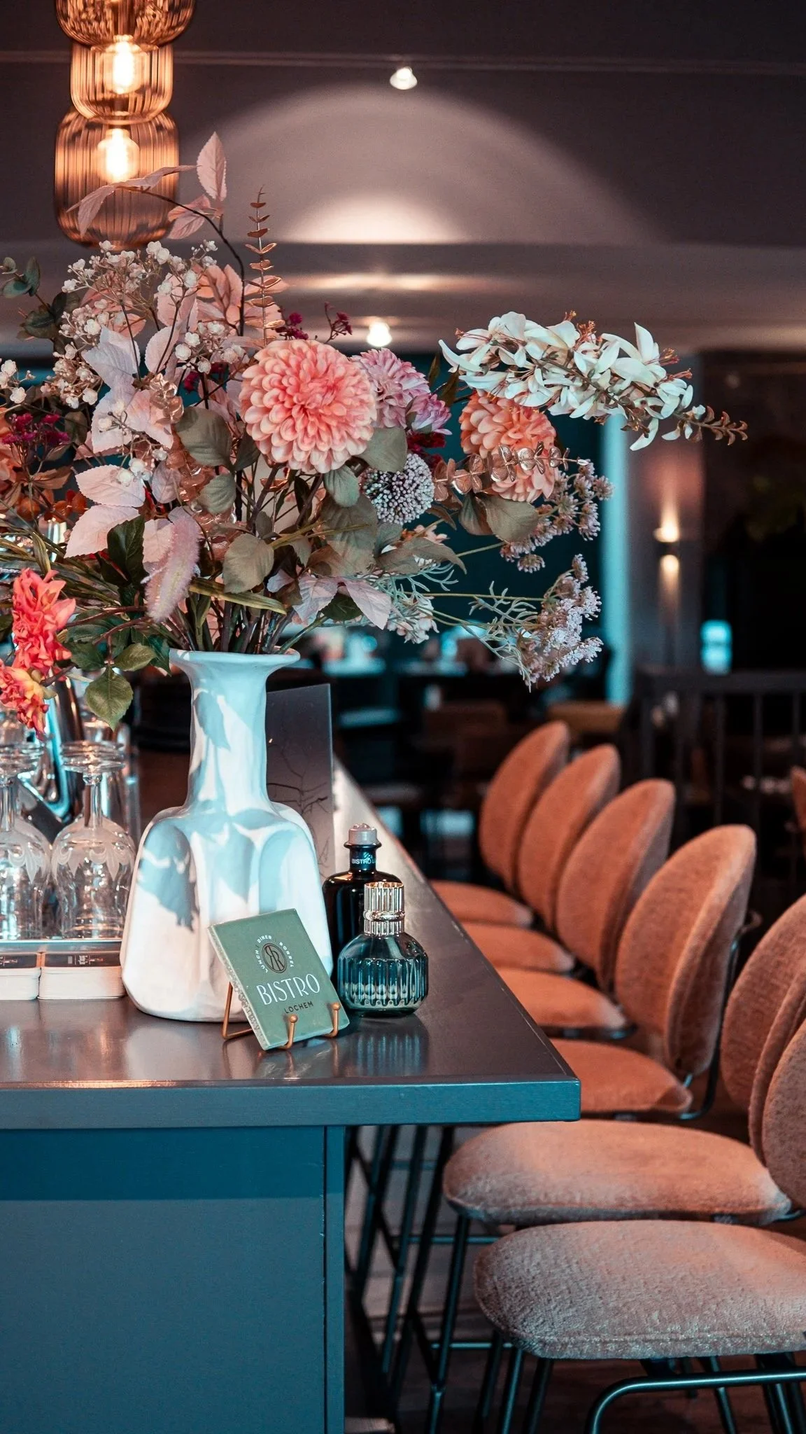 Restaurant bar with a large vase of pink flowers, upholstered stools and warm pendant lighting.
