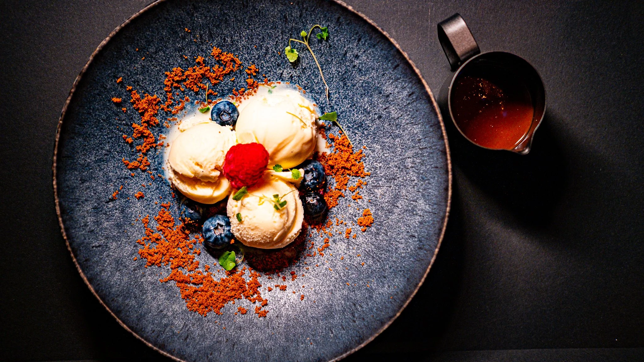 Overhead view of three scoops of ice cream with berries and crumbs on a dark plate, beside a small jug of sauce.