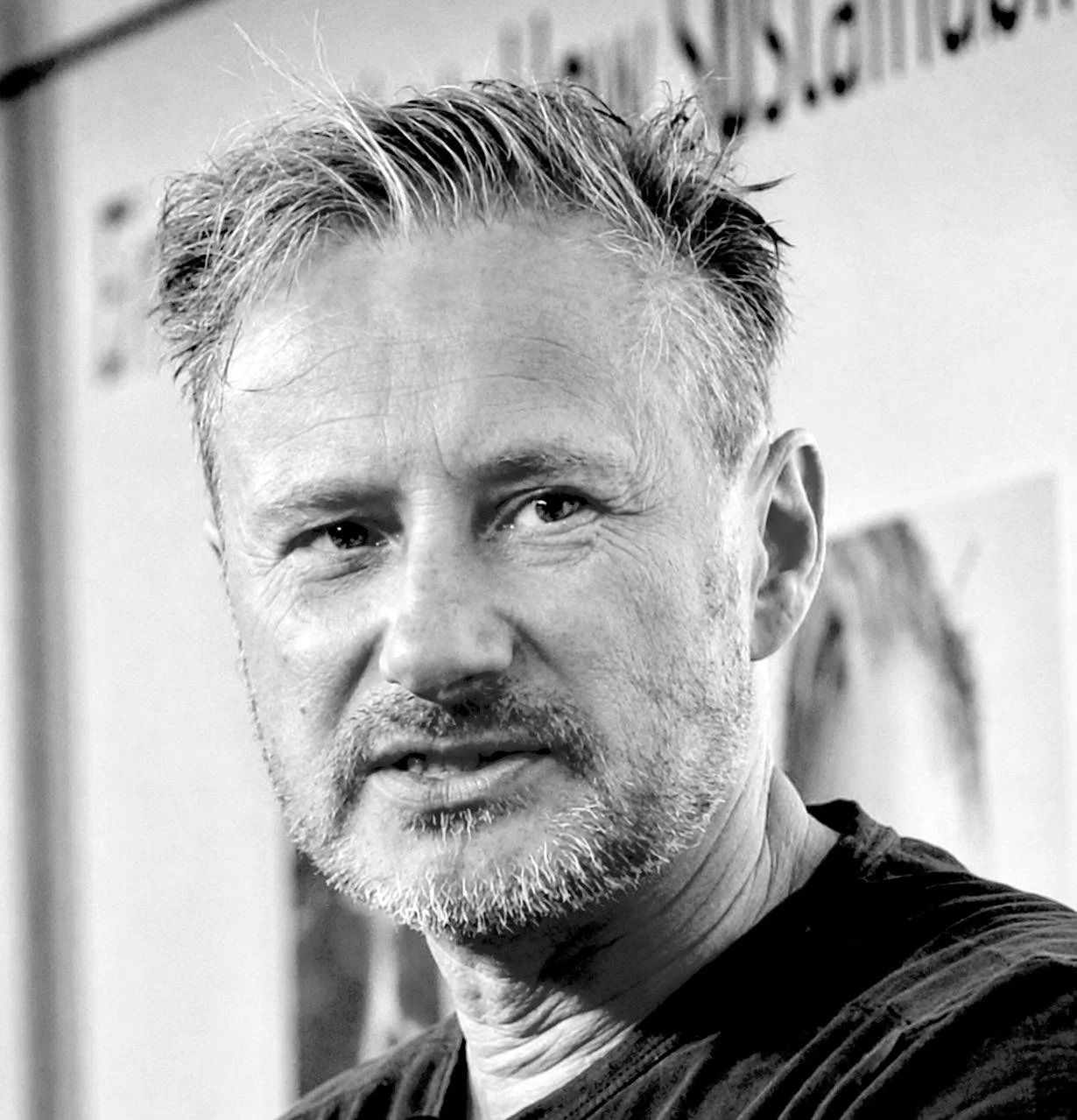 Close-up black and white photo of a middle-aged man with a beard and mustache, smiling slightly, resting his chin on his hand.