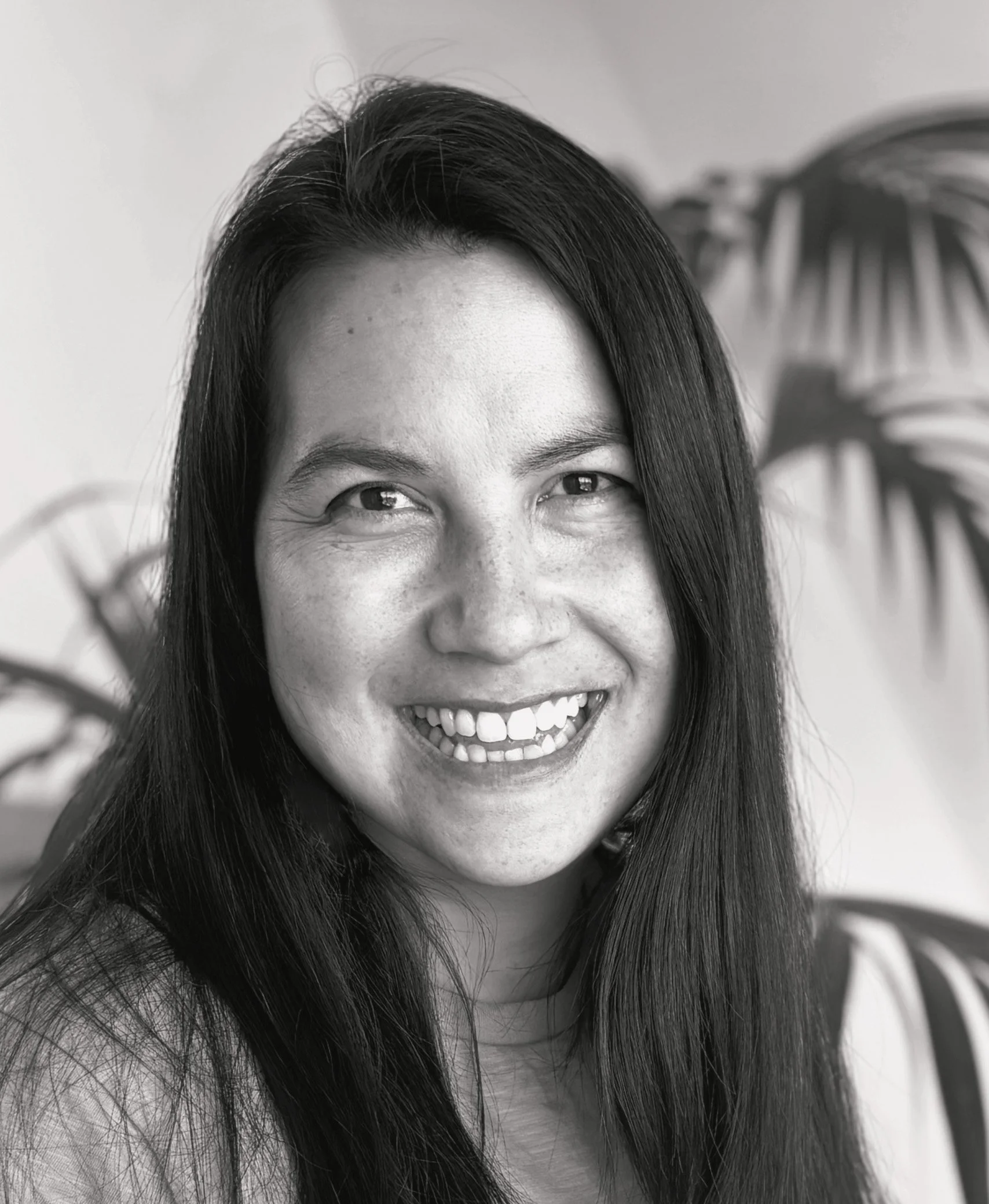 A woman with long dark hair smiling, with a background of houseplant leaves.