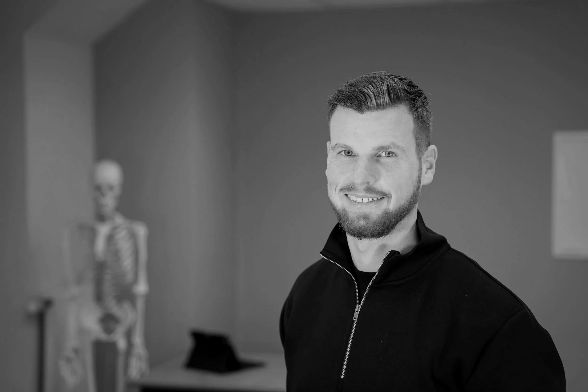 Ein junger Mann mit braunem Haar und Bart lächelt in die Kamera, im Hintergrund eine Skelett-Dekoration, das Bild ist grau-skaliert.