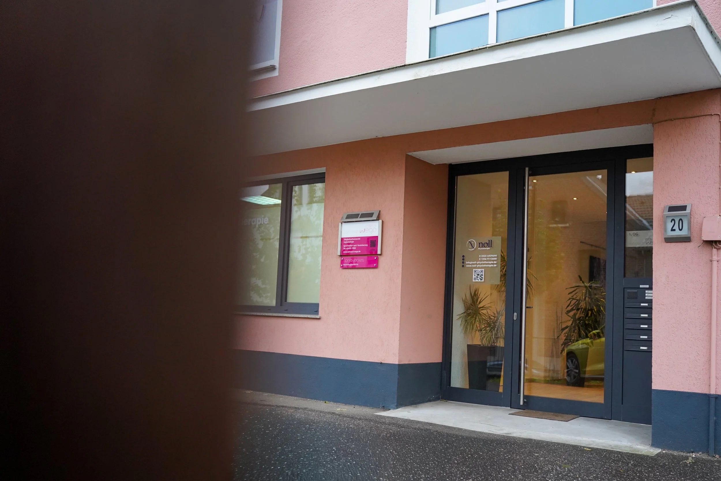 Blick auf das Eingangsportal eines Gebäudes mit Hausnummer 20, gefasst in einem schwarzen Rahmen mit Glastür, Pflanzen im Innenraum, pinkfarbige Briefkästen an der Wand und eine schwarze Hausnummer. Der vordere Bereich ist teilweise durch eine dunkle