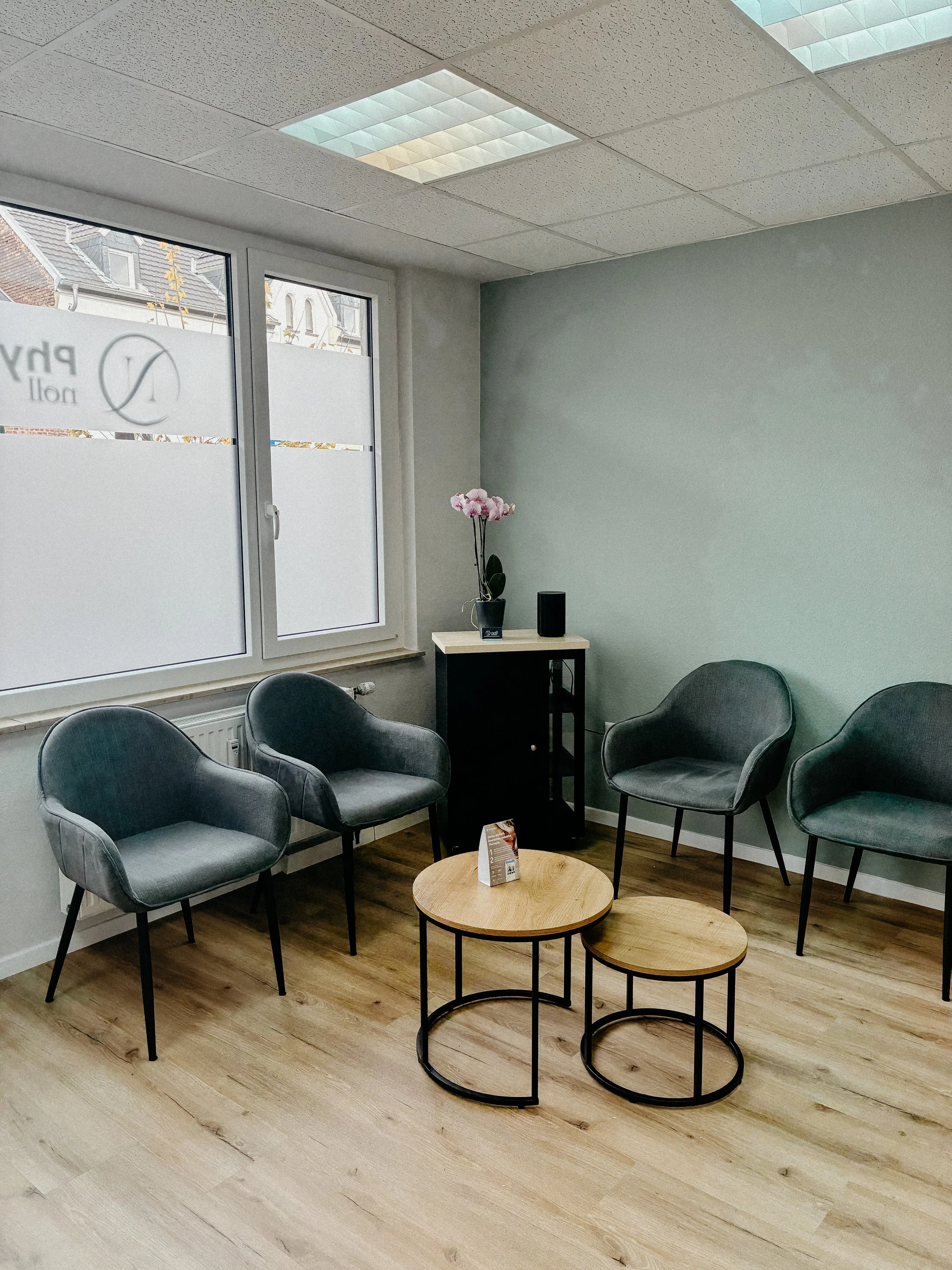 Warteschleifenbereich mit vier grauen Sesseln, zwei runden Holz-Couchtischen, einer kleinen schwarzen Ablage mit einer Orchidee und einem Lautsprecher, große Fenster mit frosted Glas, weiße Wände und Holzboden.